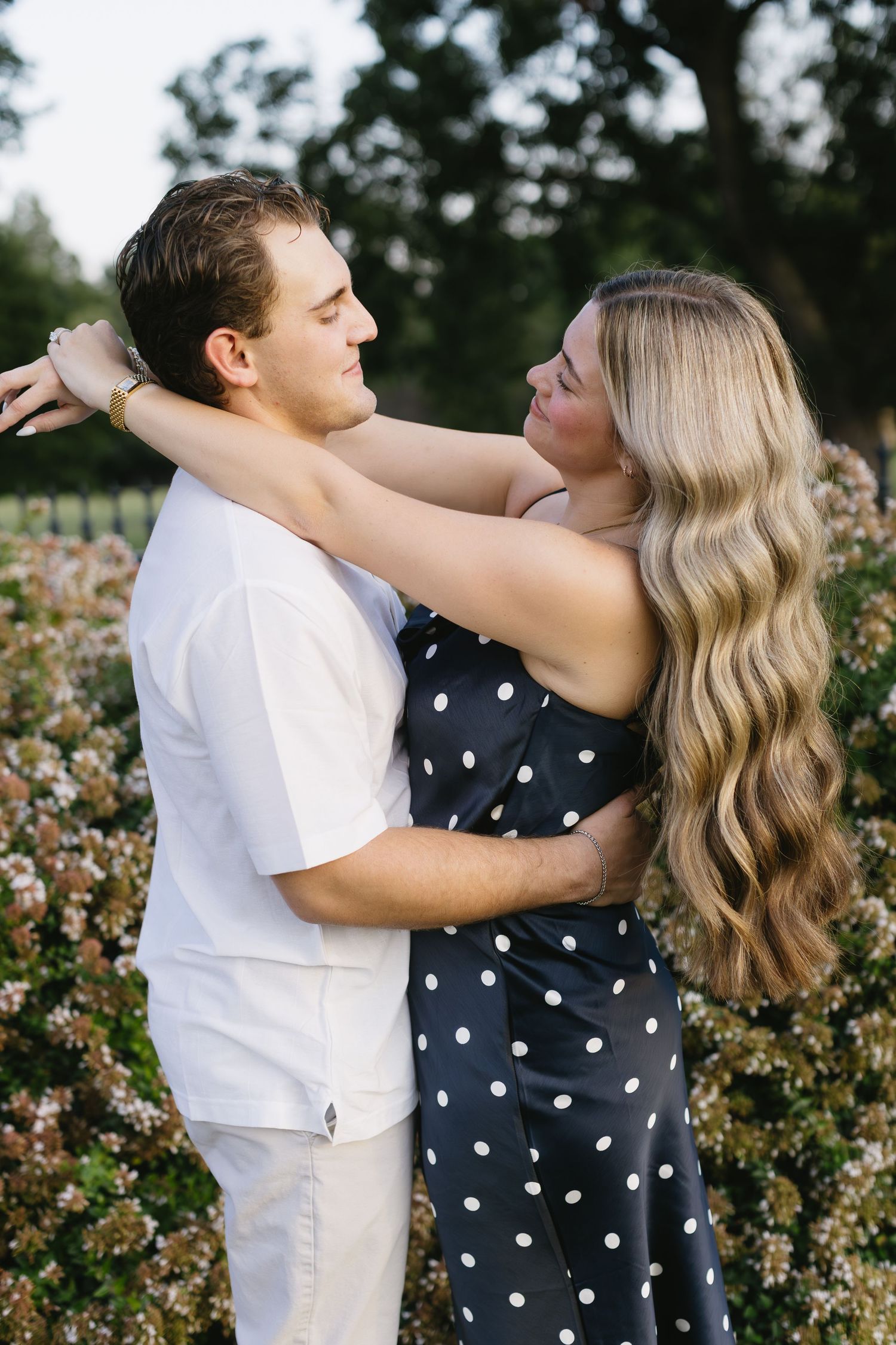 A romantic couple embraces near blooming rose bushes, with her wearing a polka dot dress and him in white.