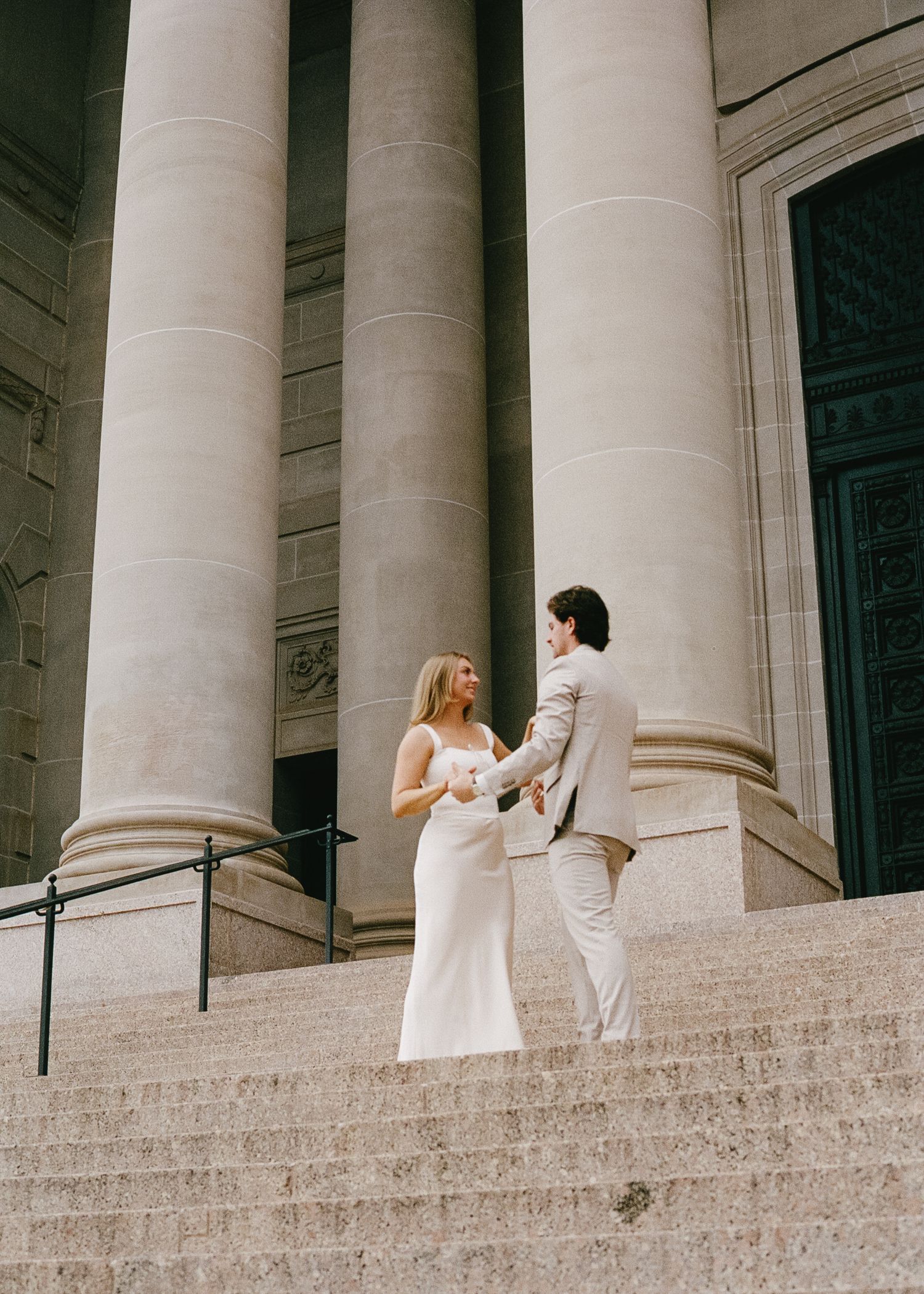 A couple in formal attire stands on grand stone steps with classical columns in the background.