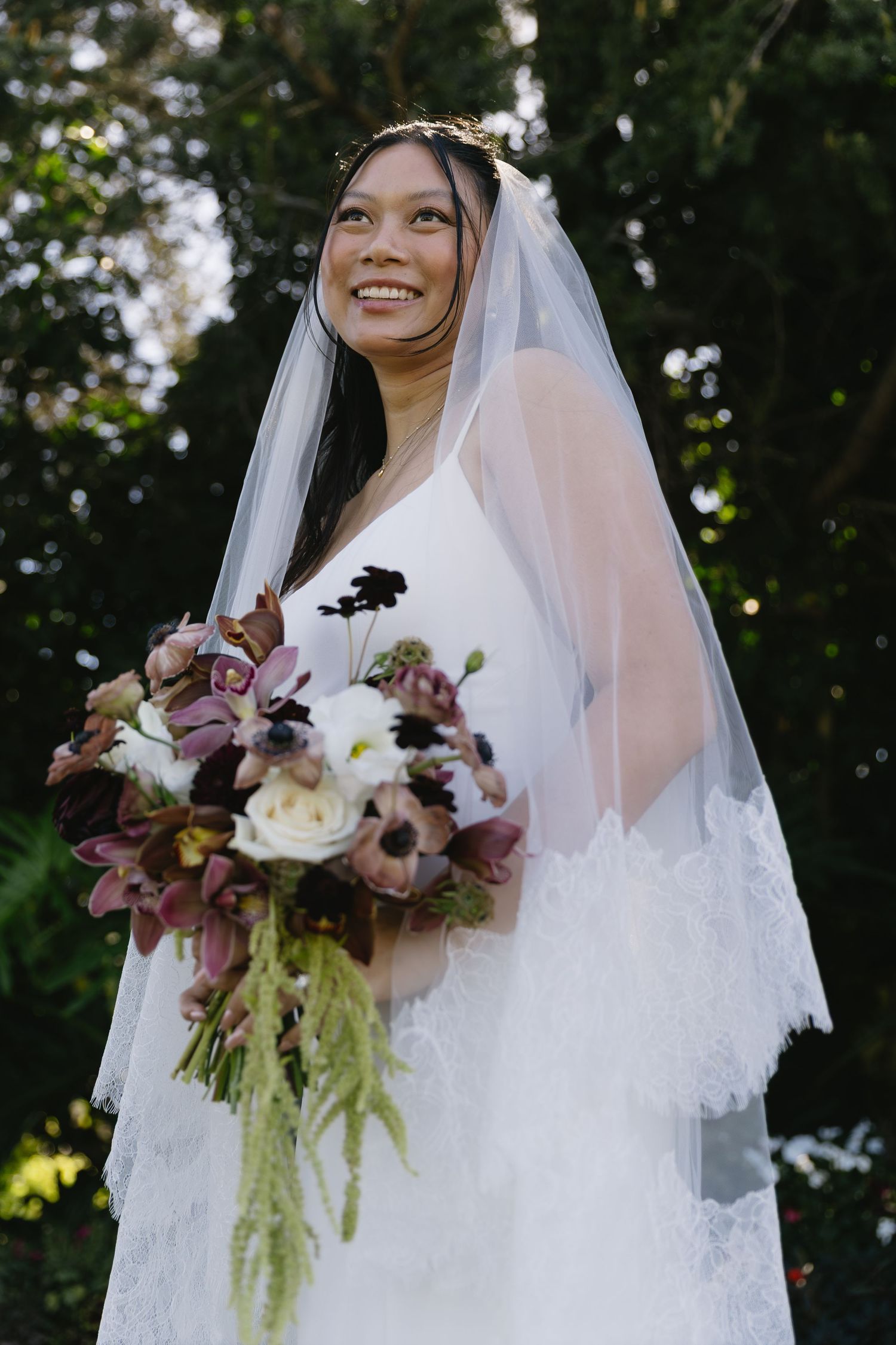 Bride in white lace dress holds elegant purple and white floral bouquet.