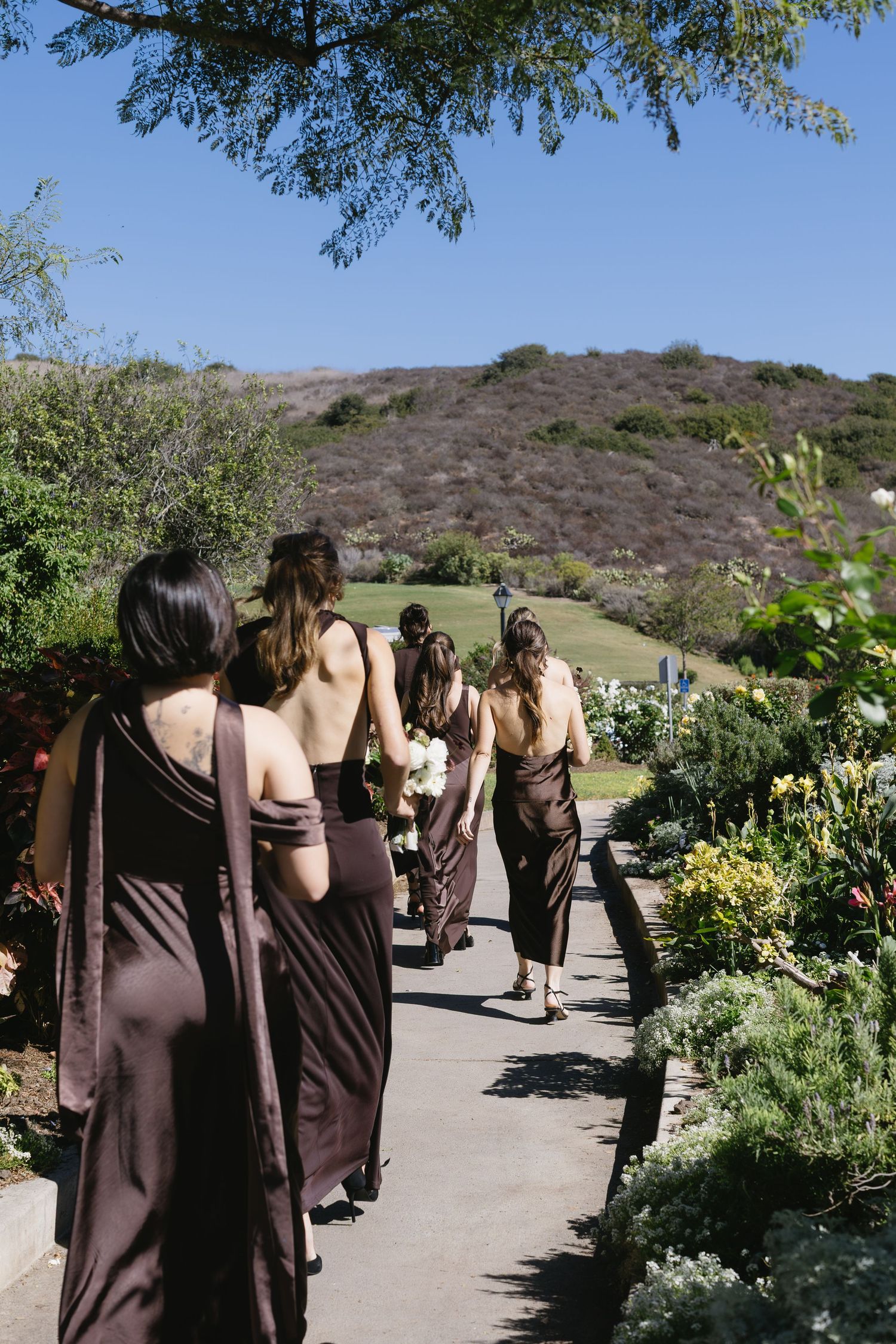 Bridesmaids in brown dresses walking along a garden path with mountains visible in the background.