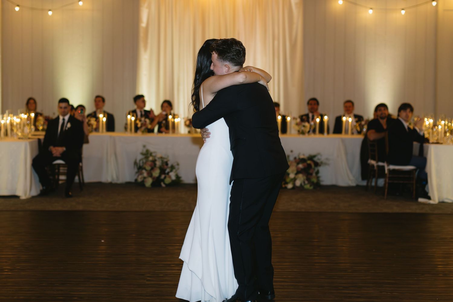 A couple shares their first dance in an elegant reception hall with white draping and ambient lighting.