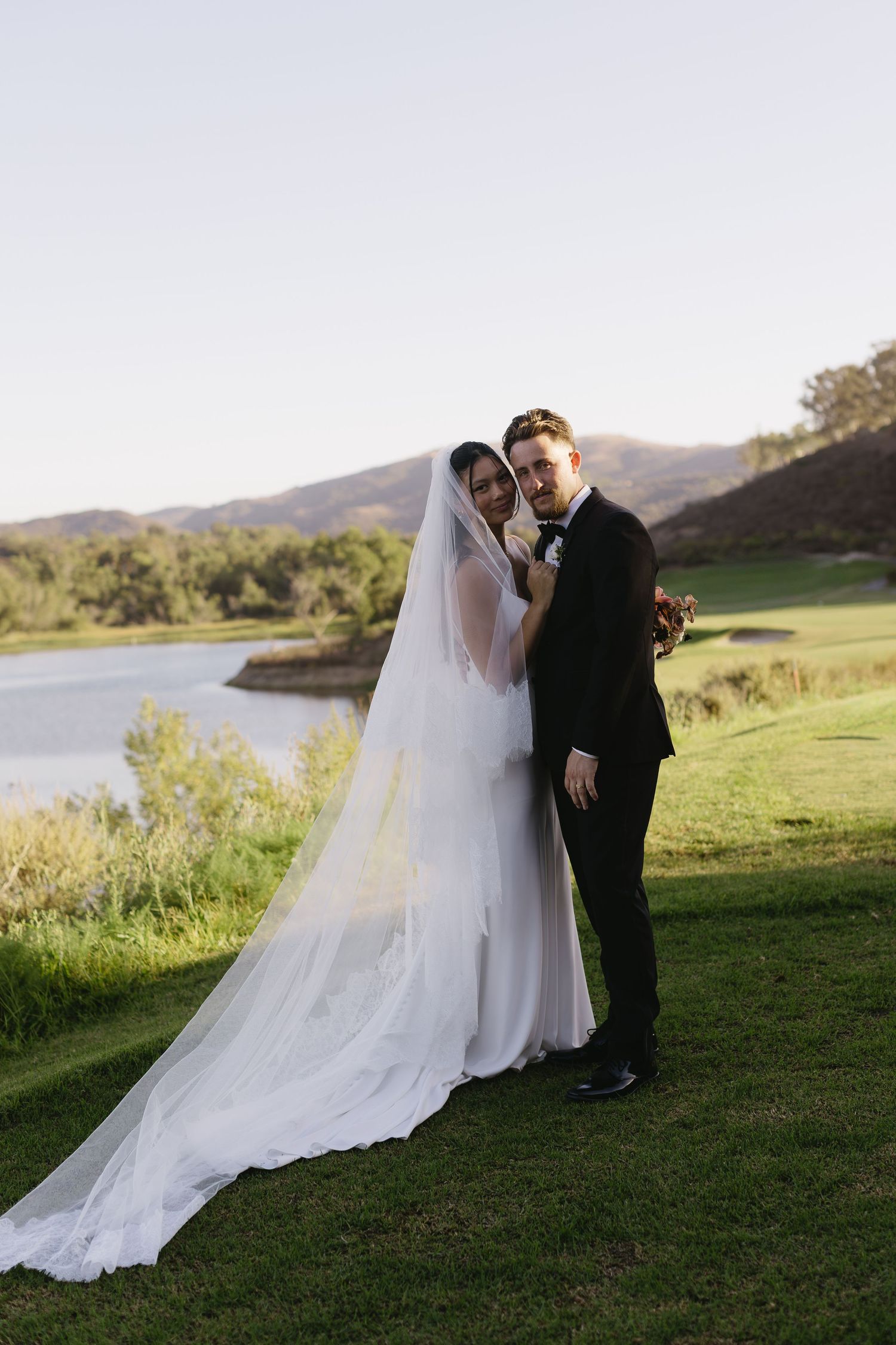 A romantic wedding couple poses on a grassy hillside overlooking a serene lake during golden hour.