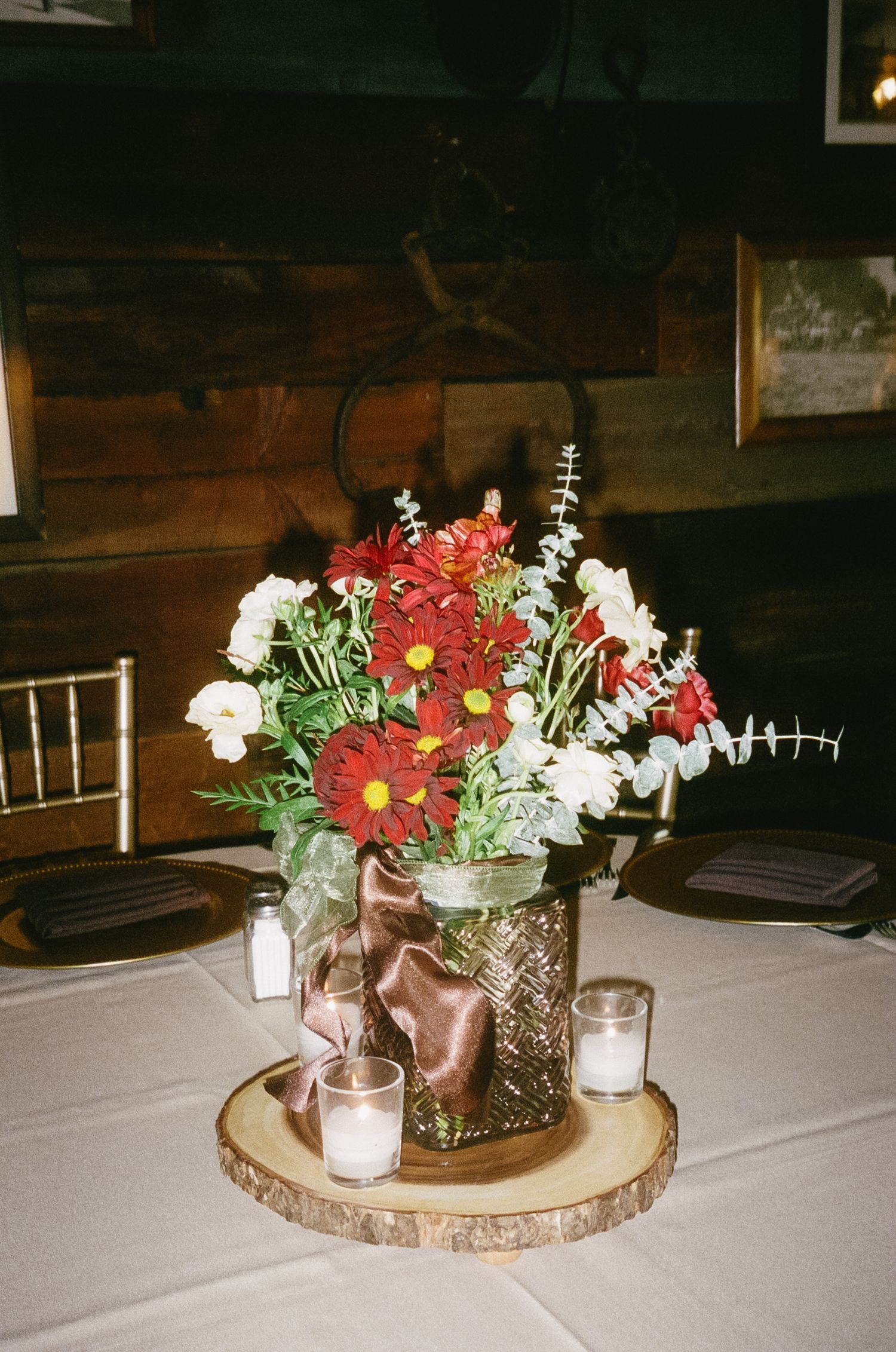 A rustic wedding centerpiece with red and white flowers in a glass vase on a wooden slice base with candles.