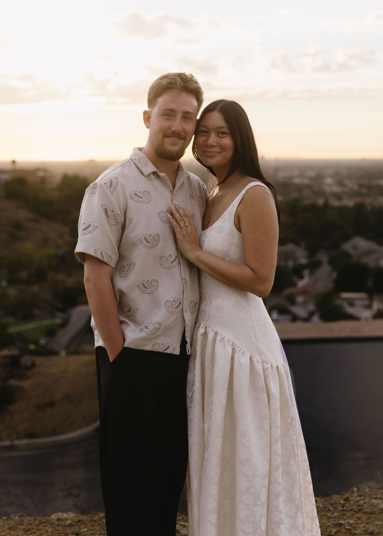 A couple embraces on a hilltop overlook at sunset, with the woman wearing a white dress and man in a patterned shirt.