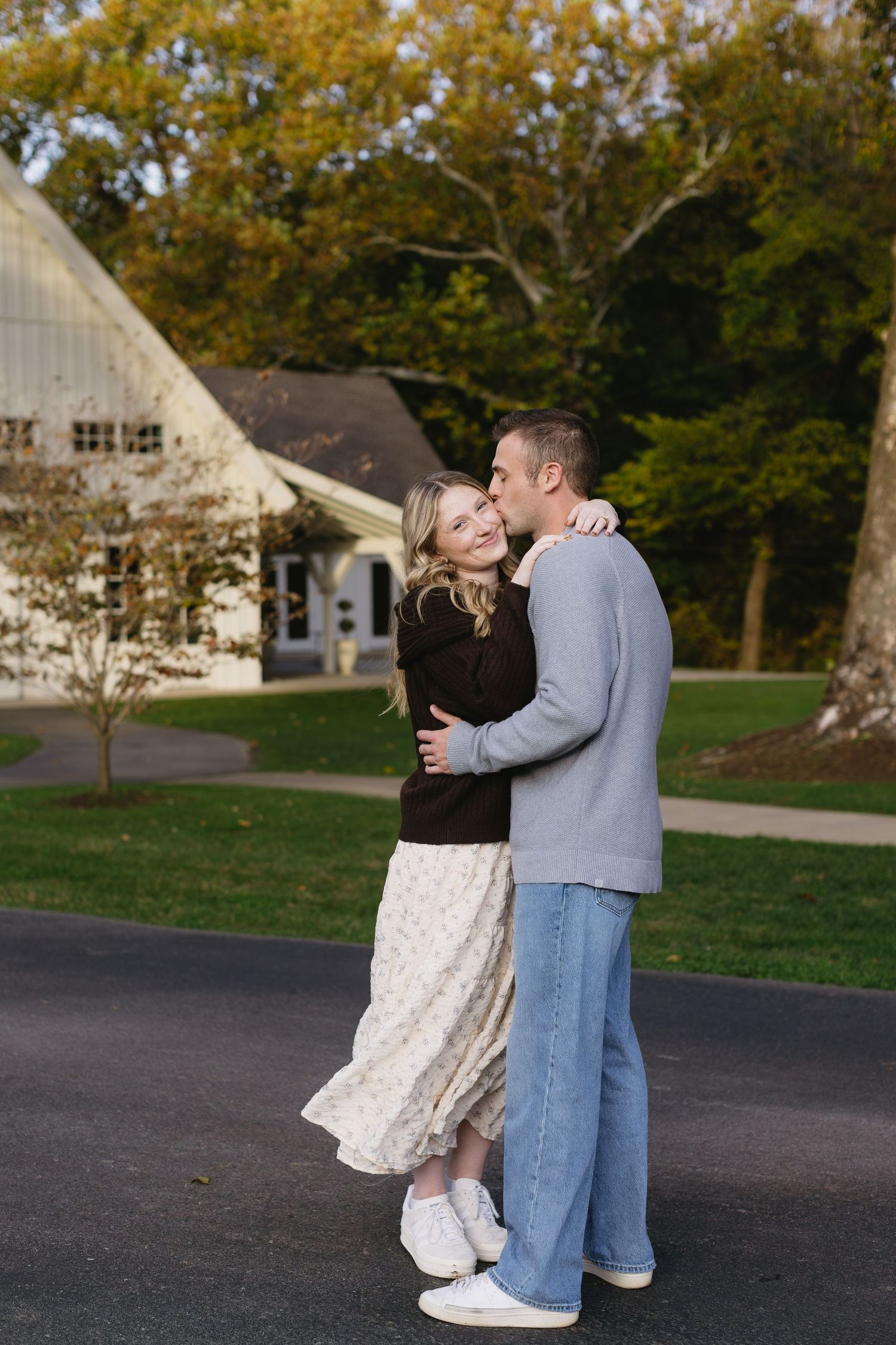 A couple embraces while standing on a paved path with autumn trees and a house in the background.