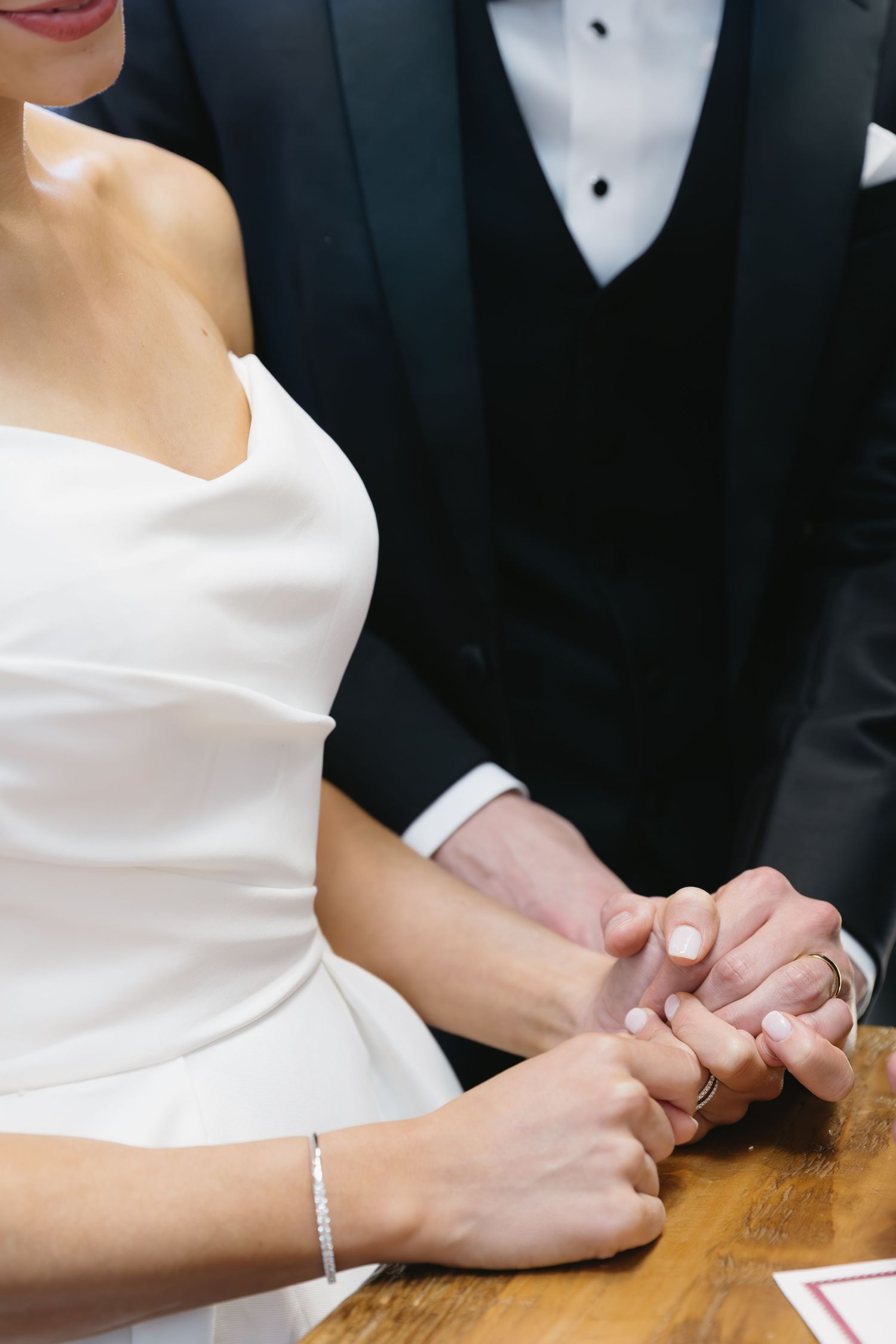 Close-up of newlyweds holding hands at a table, showing white wedding dress and black tuxedo details.