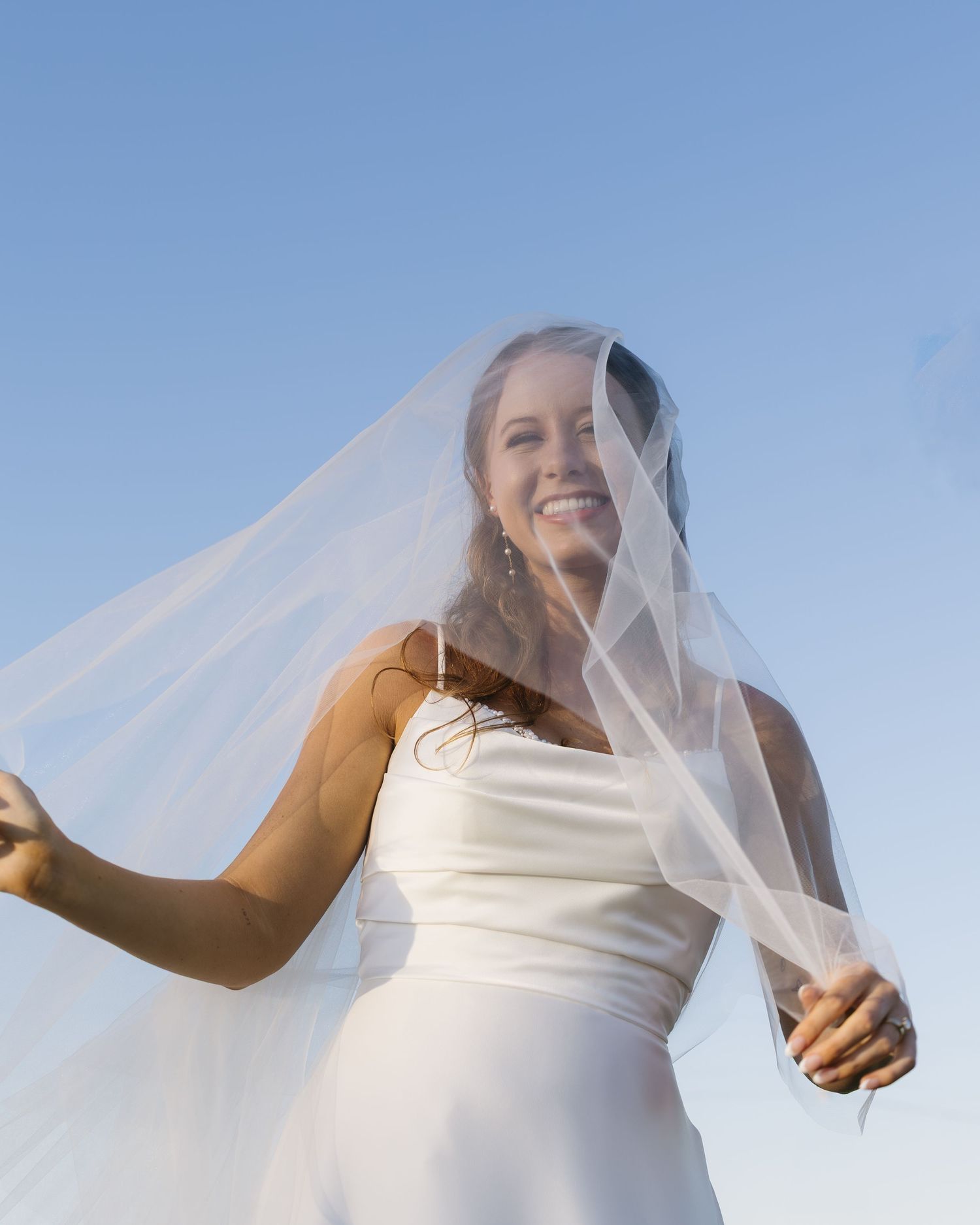 Sunlit scene of flowing white wedding veil billowing against bright blue sky during outdoor bridal photoshoot.