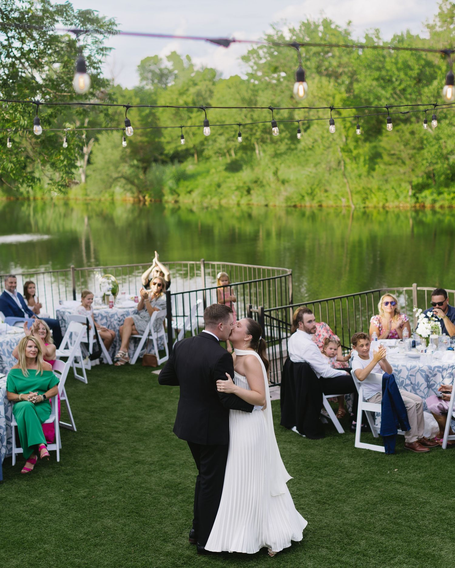 Outdoor wedding reception by a lake with white chairs and dancing guests.