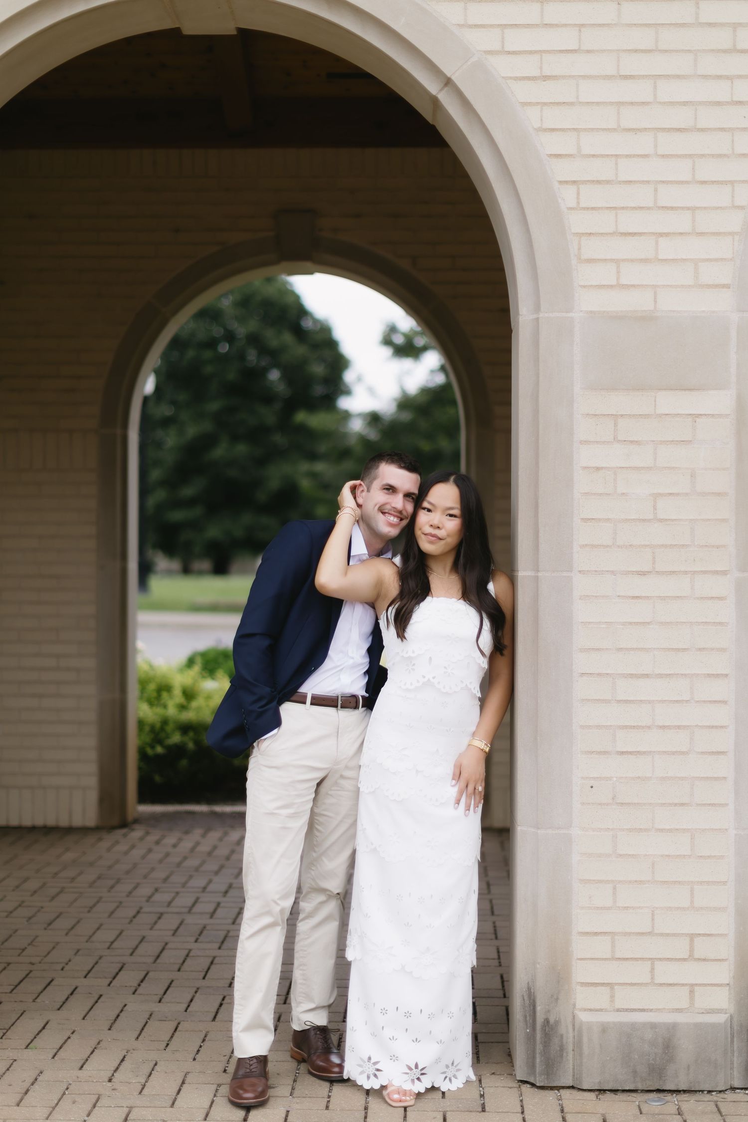 A loving couple shares a romantic moment under a white brick archway in an outdoor setting.