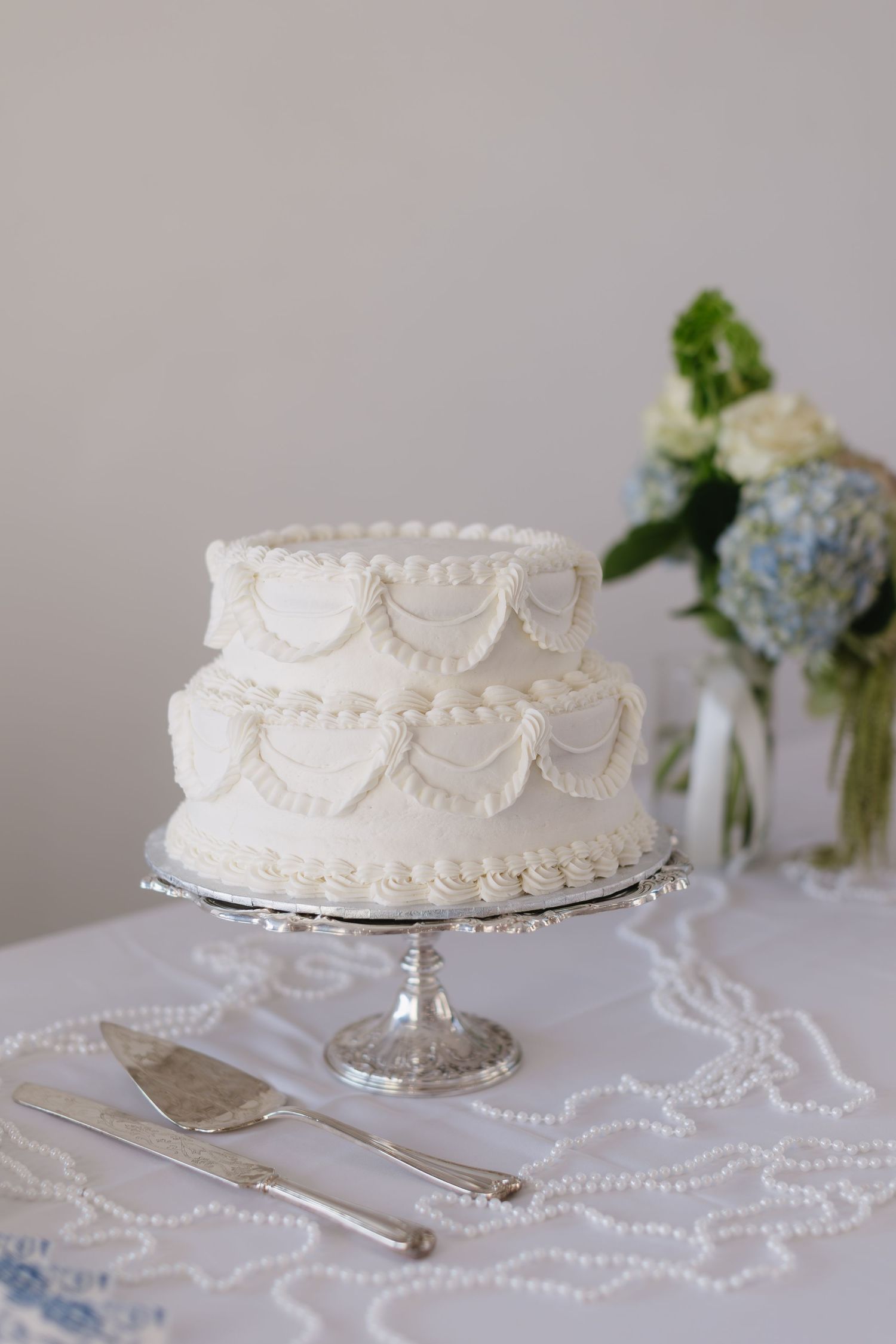 White wedding cake with delicate piping details displayed on a silver cake stand with pearl decorations.