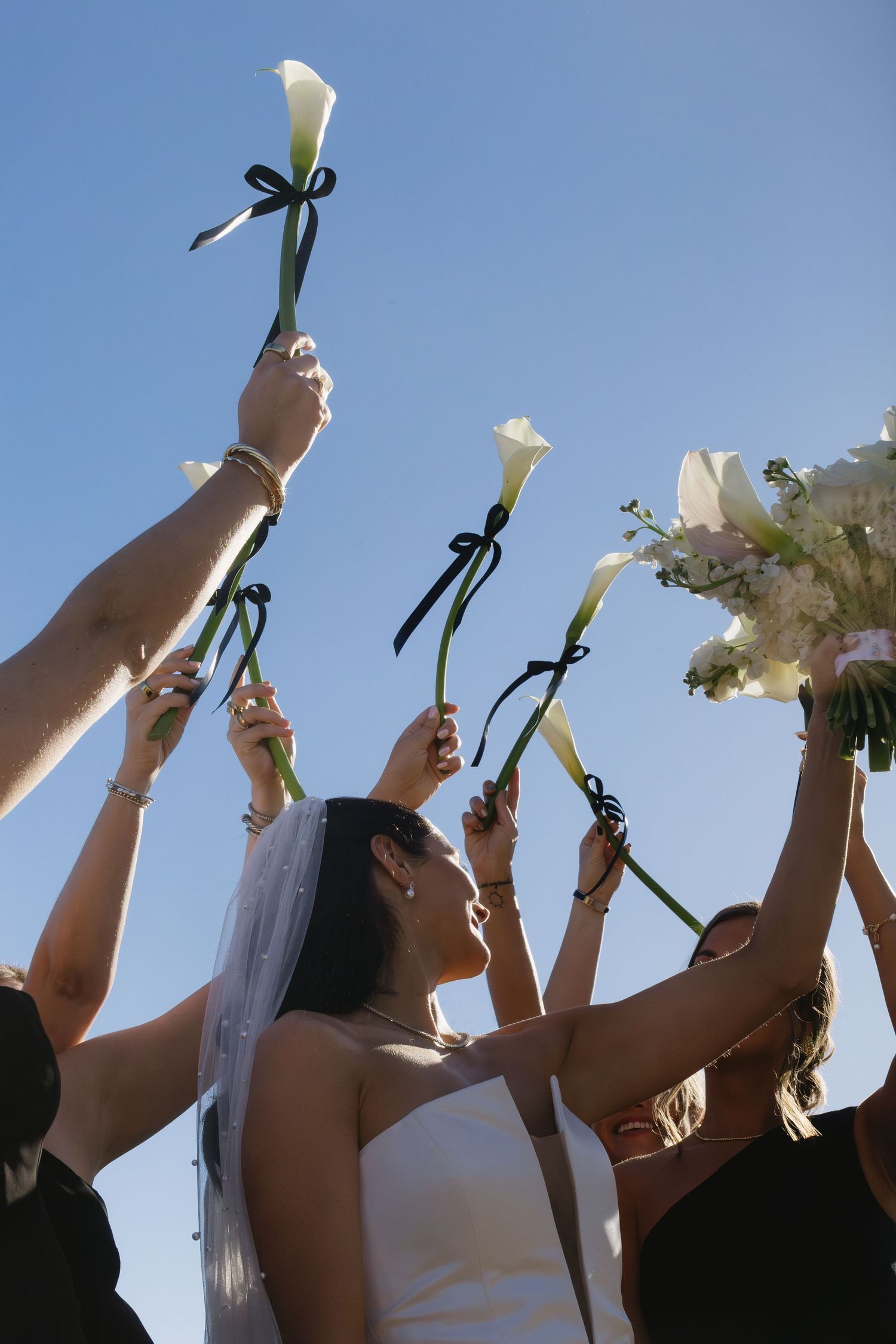 A group of people holding white calla lily flowers up towards a bright blue sky in a celebratory gesture.