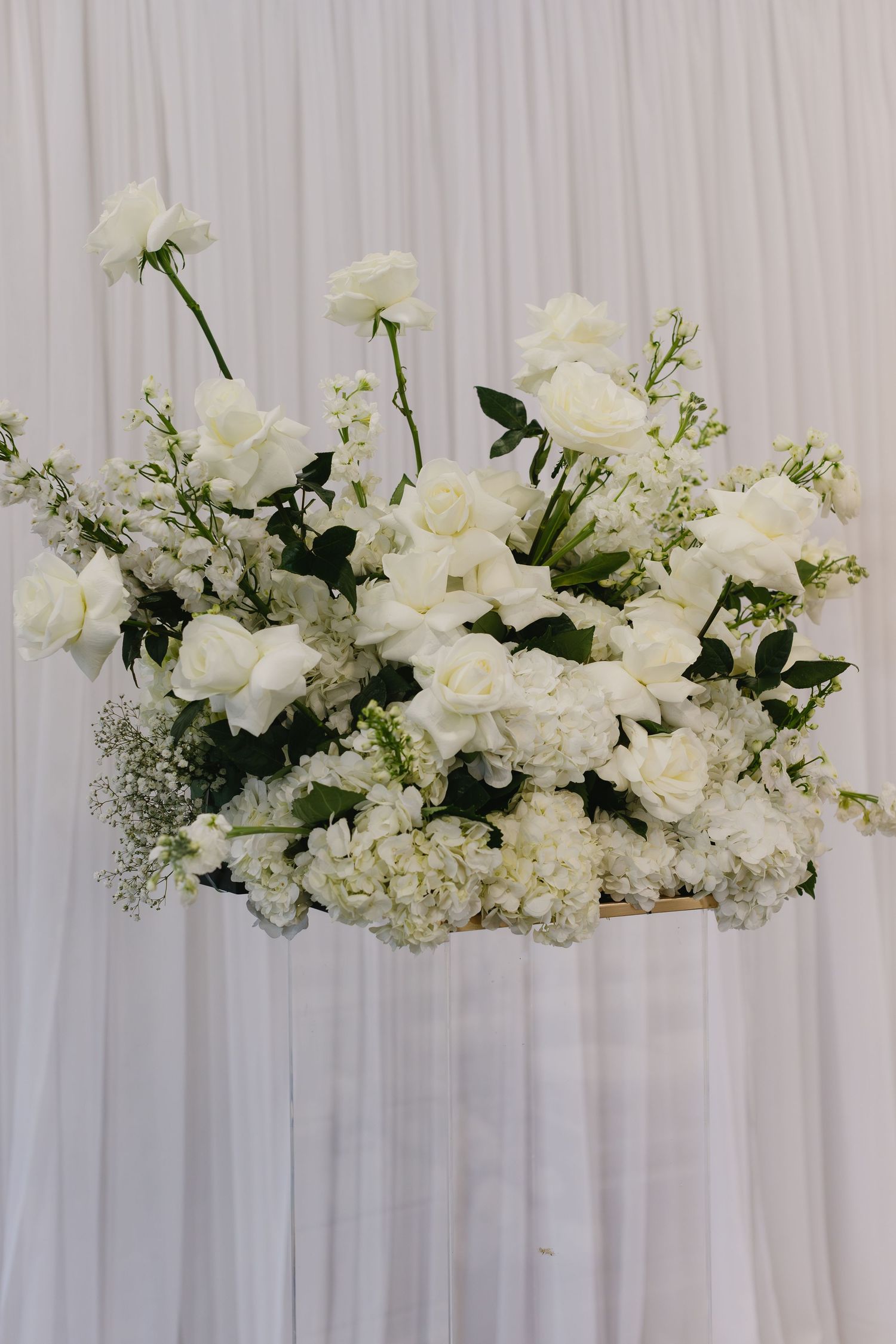 Elegant white floral arrangements with roses and greenery decorating a white backdrop for a wedding ceremony.