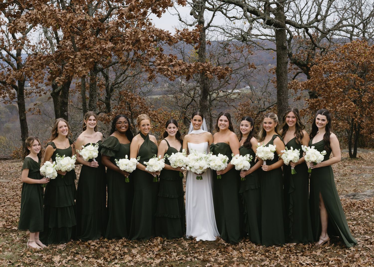 A wedding party in dark green dresses poses with the bride in white among autumn trees.