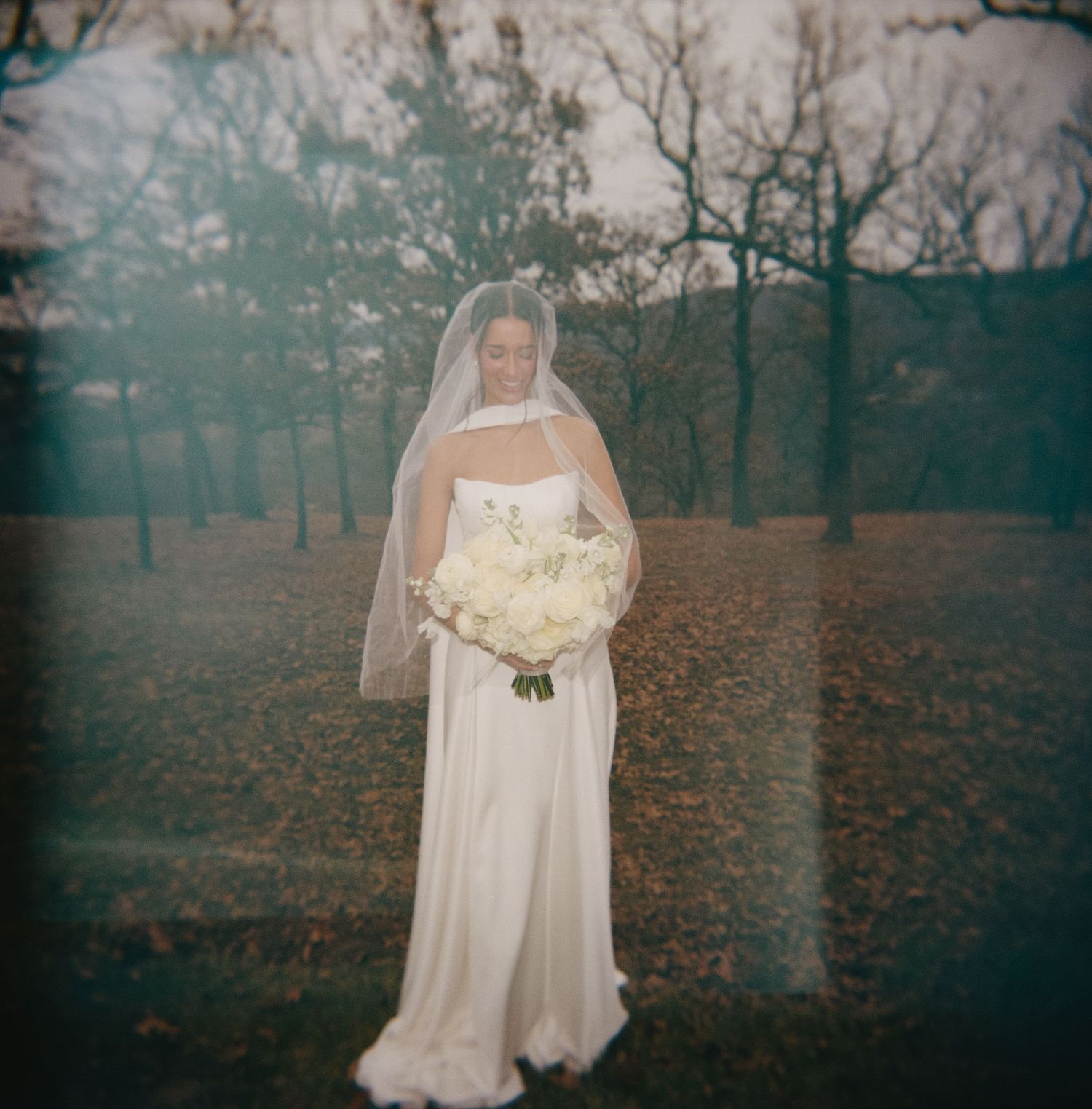 A bride in a white wedding dress poses in a misty autumn forest setting with fallen leaves on the ground.