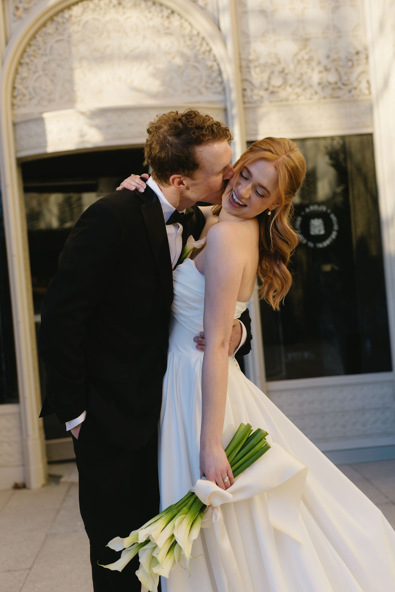 Wedding couple shares a joyful moment with white calla lily bouquet against stone architecture.