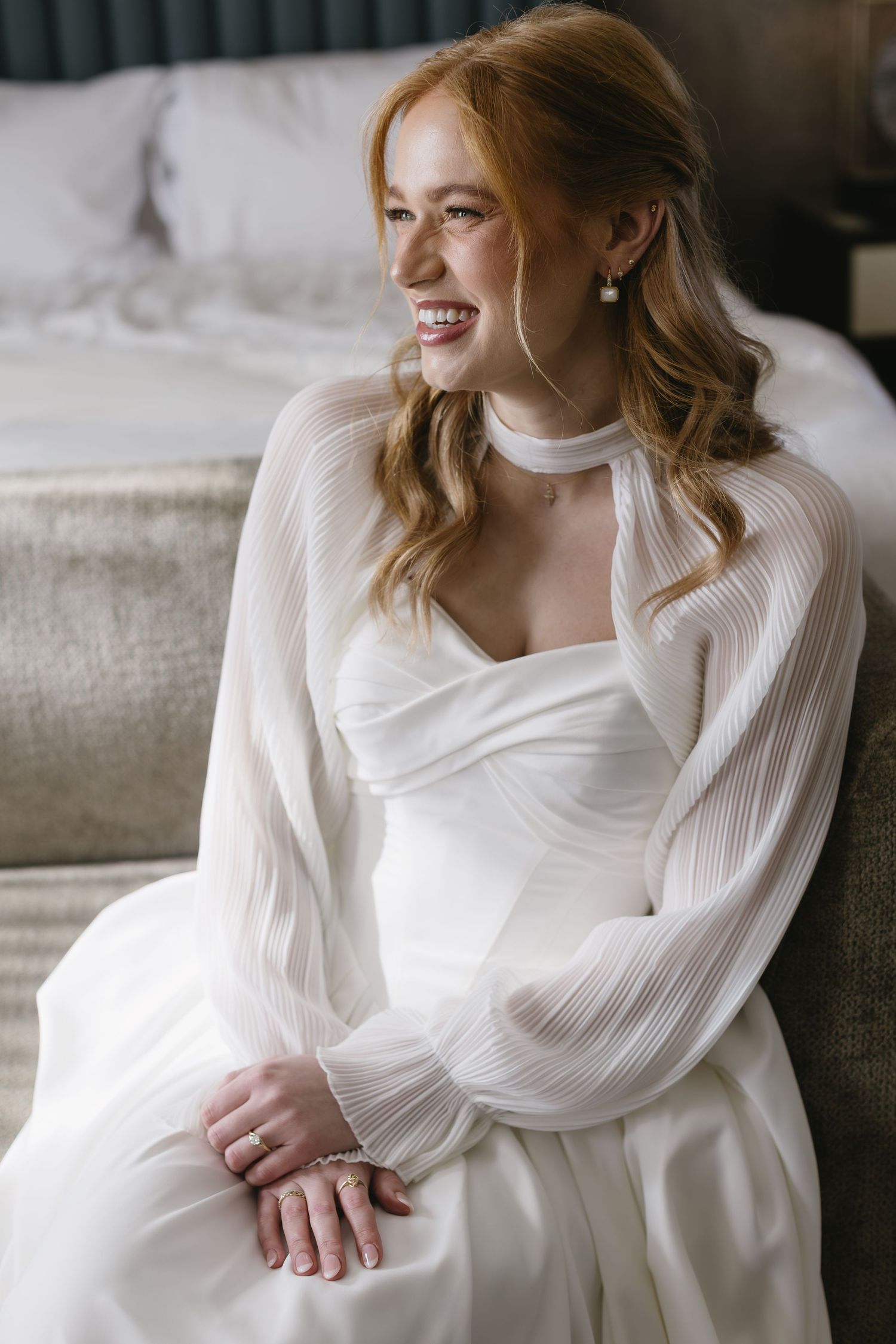 A bride in a white dress with sheer sleeves sits elegantly on a couch.