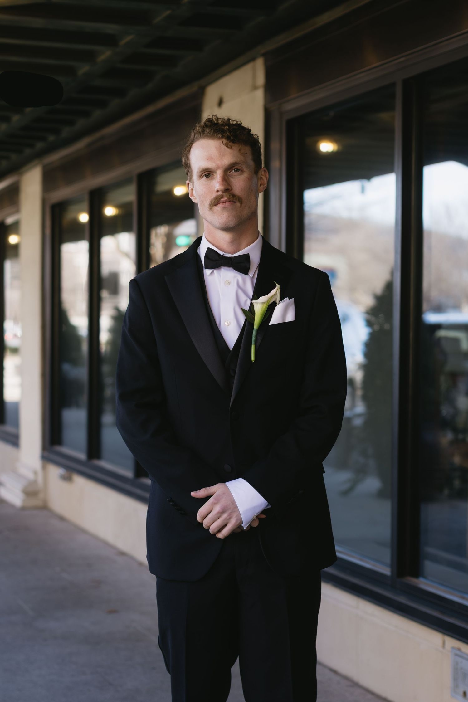 Groom in classic black tuxedo and bowtie photographed from different angles in an outdoor corridor.