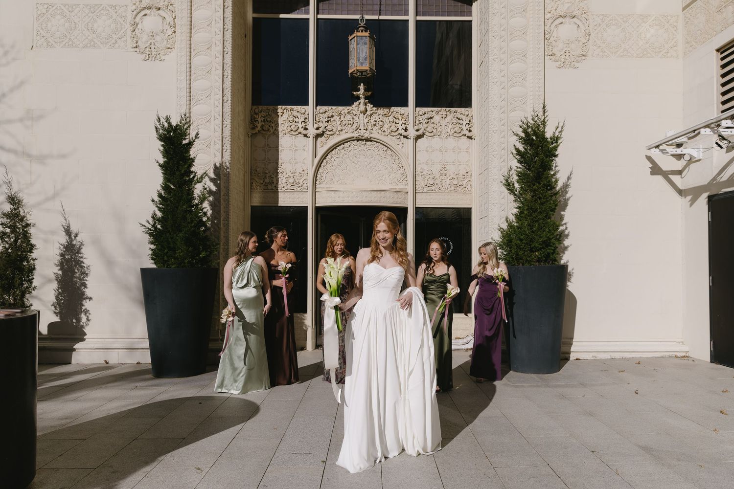 Wedding party in white dresses and purple gowns pose elegantly against a light stone building exterior.