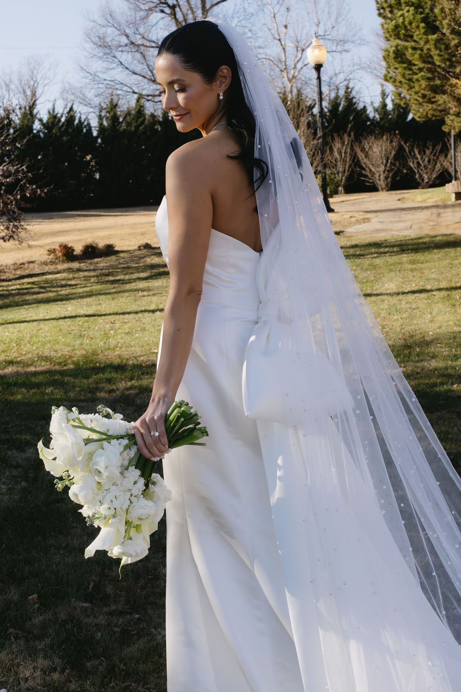 Bride in elegant white gown holds orchid bouquet outdoors during golden hour.