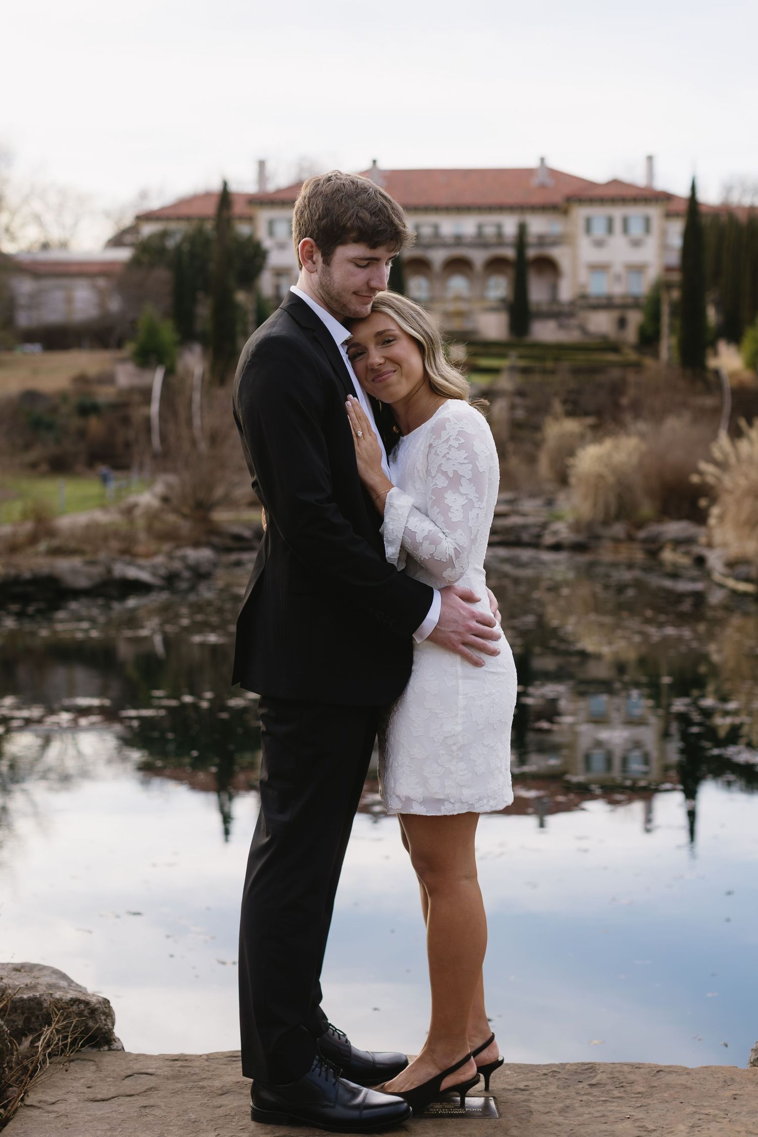 A couple shares a tender moment by a lake with Mediterranean-style architecture in the background.