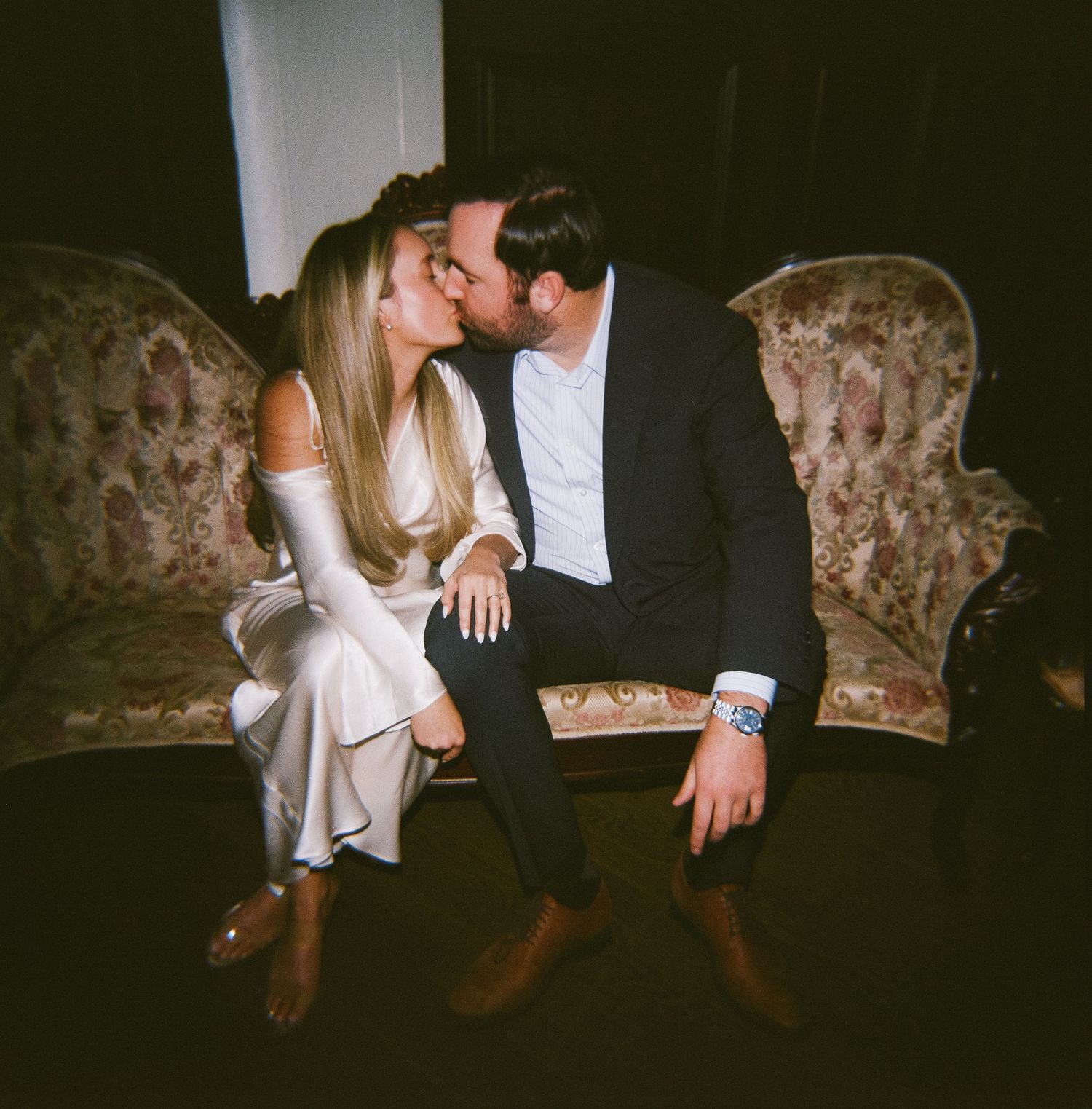 Romantic couple sitting together on vintage floral upholstered furniture in moody lighting.