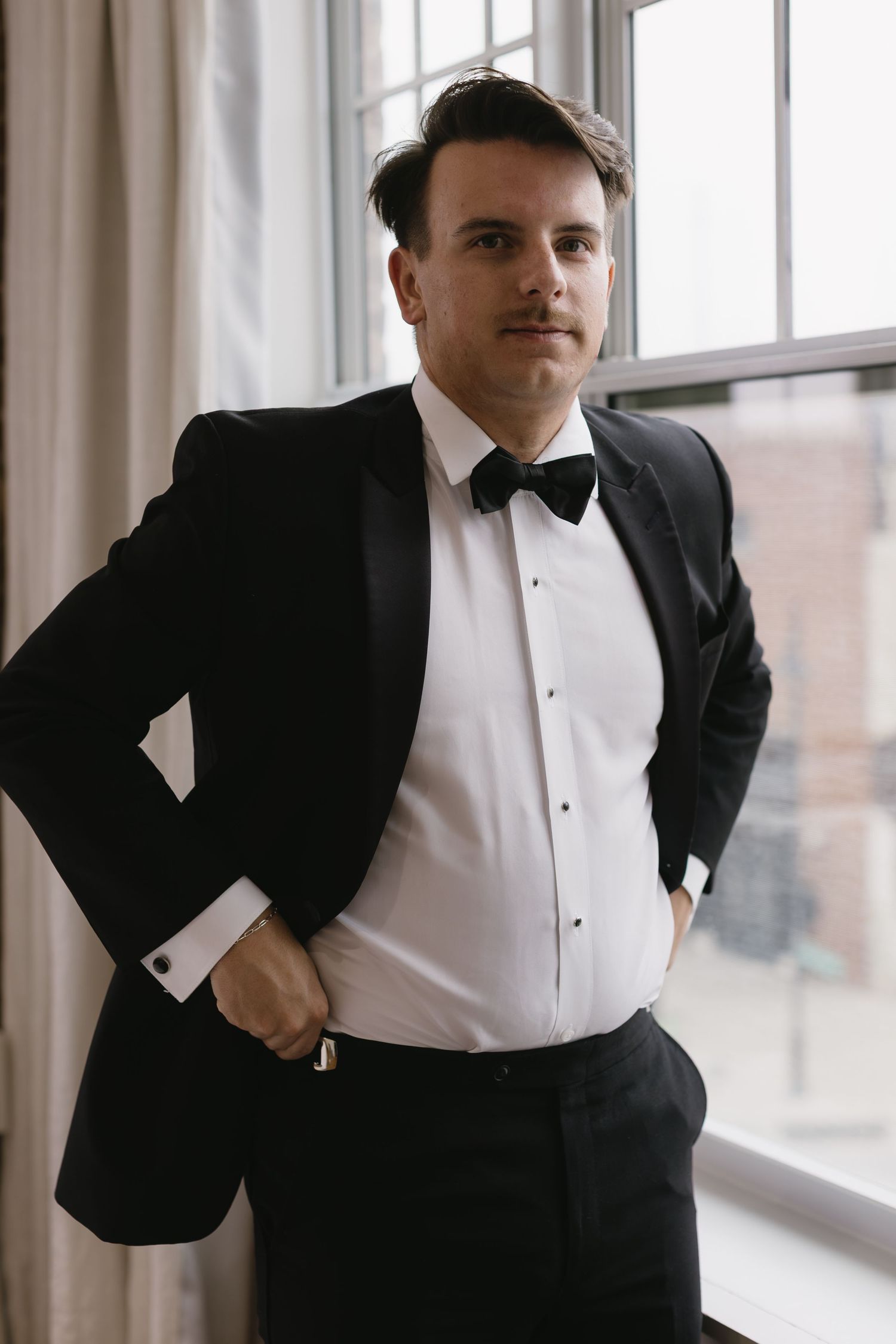 A series of four photos showing a groom in a black tuxedo and white shirt posing near windows.