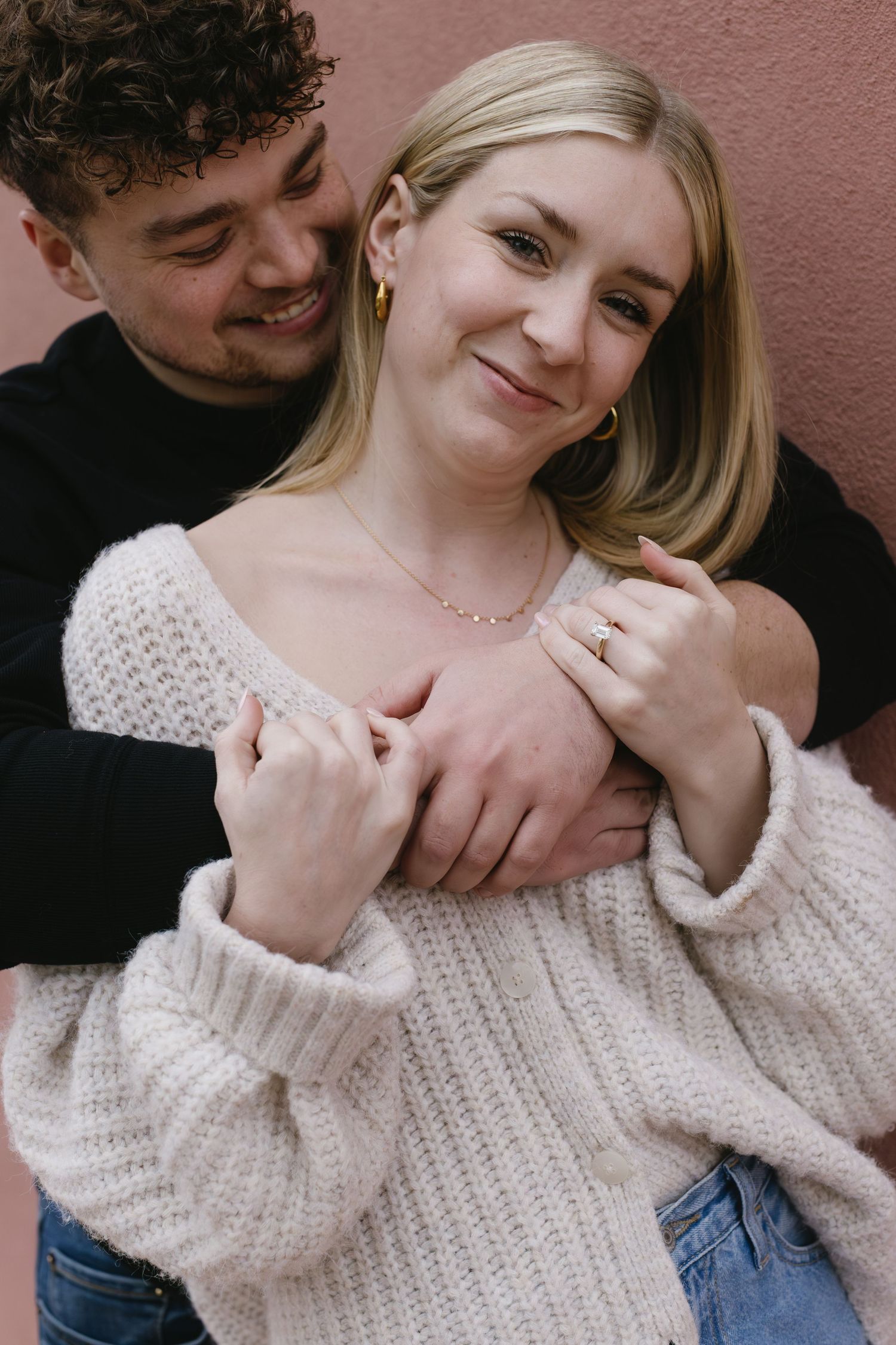 Romantic couple embracing while wearing cozy sweaters and jeans against a pink wall.
