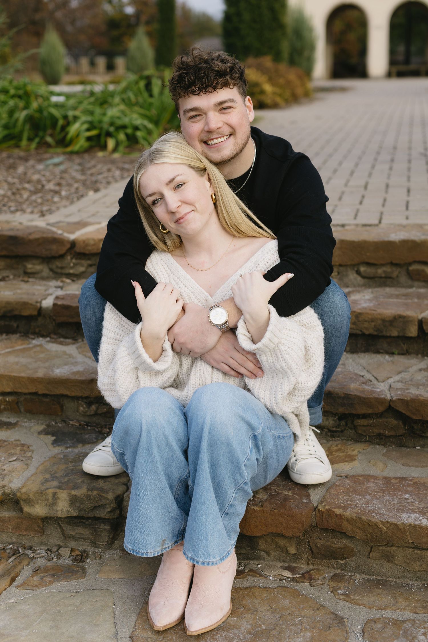 A couple sits casually on stone steps wearing jeans and sweaters in an outdoor setting.