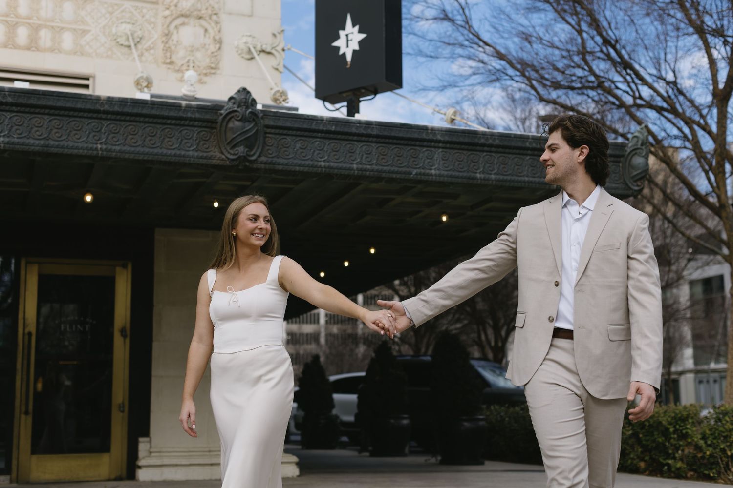 A couple in light-colored formal wear holds hands outside a building with a black banner.