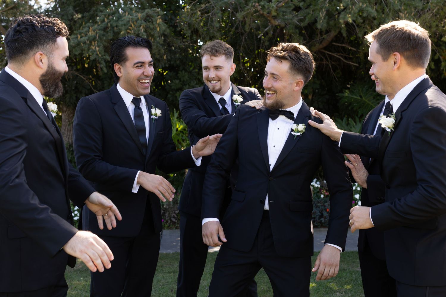 Groomsmen in black tuxedos sharing laughs during outdoor wedding photos.