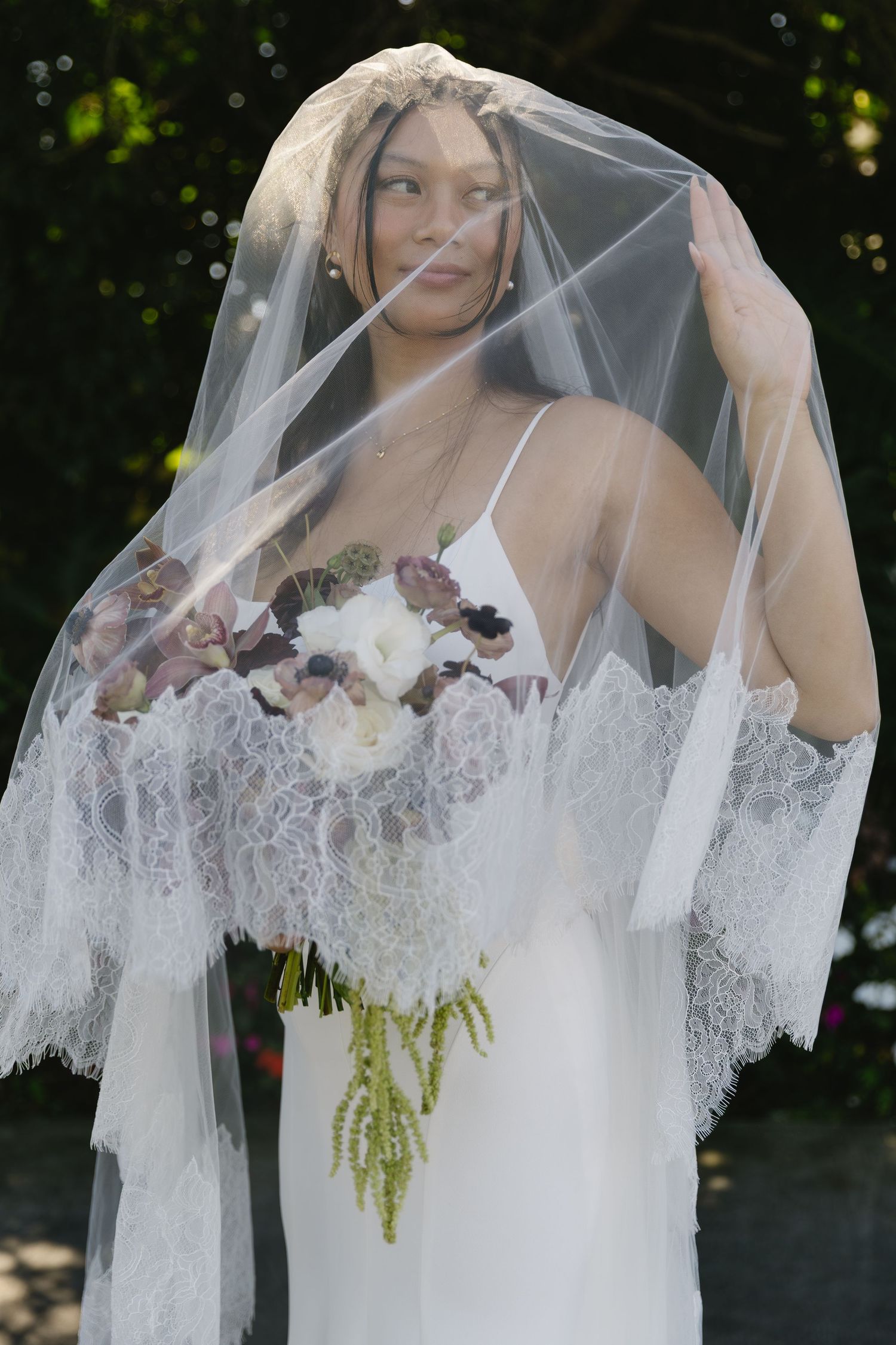 A delicate lace-trimmed veil frames a bridal portrait with cascading florals and greenery.