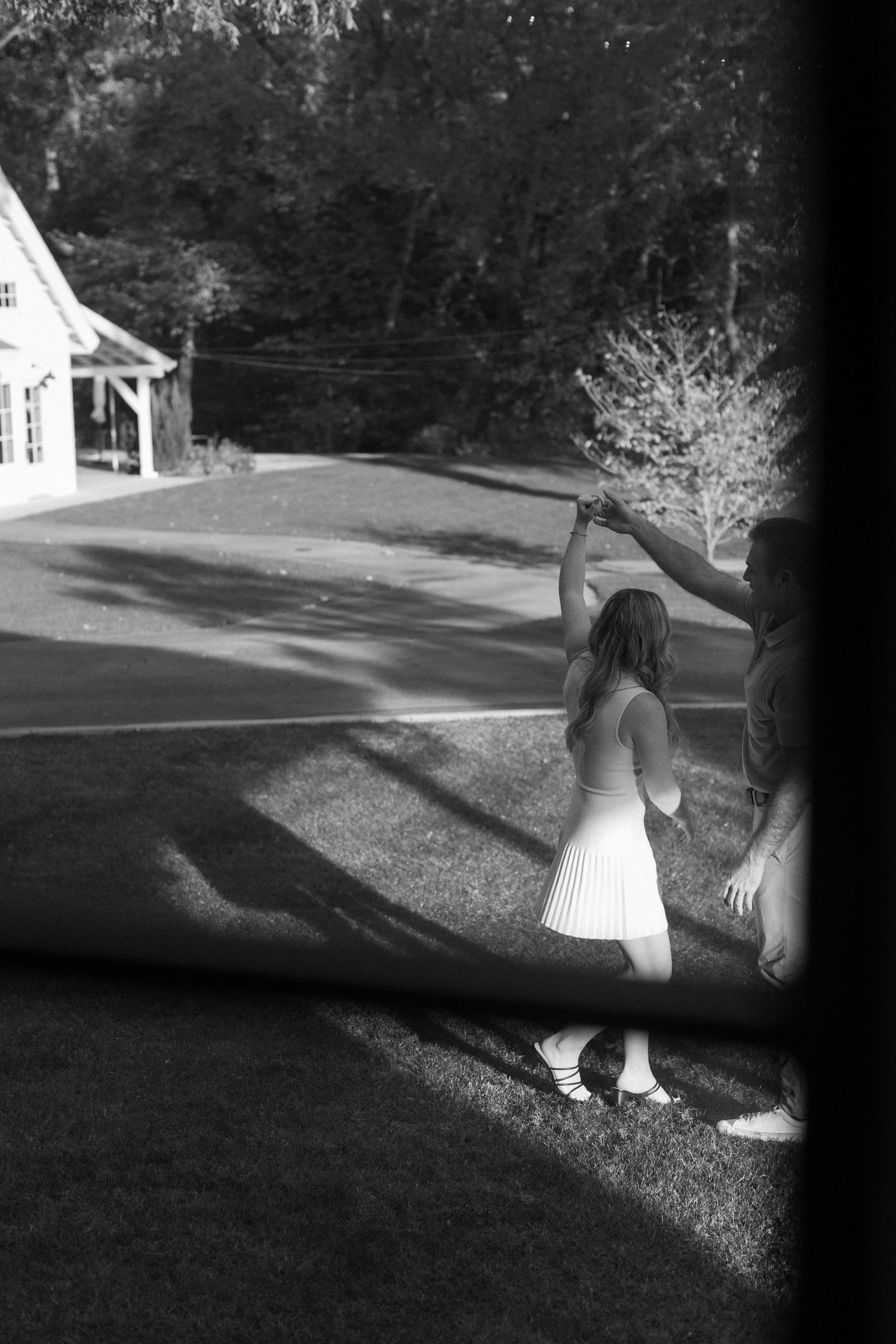 Black and white photo of someone in a white dress dancing outdoors near a house and trees casting long shadows.