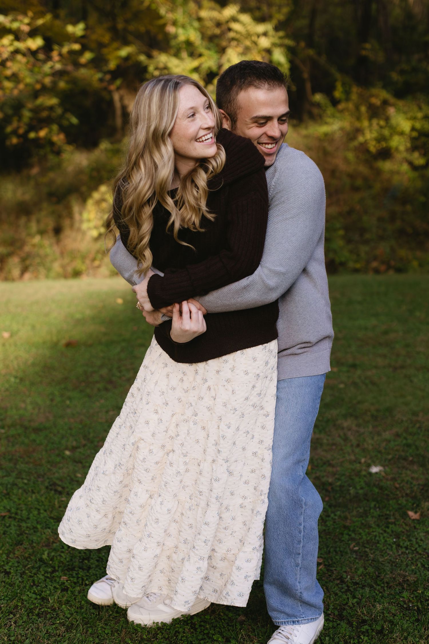 A couple laughs together while embracing outdoors on a grassy lawn with fall trees in background.