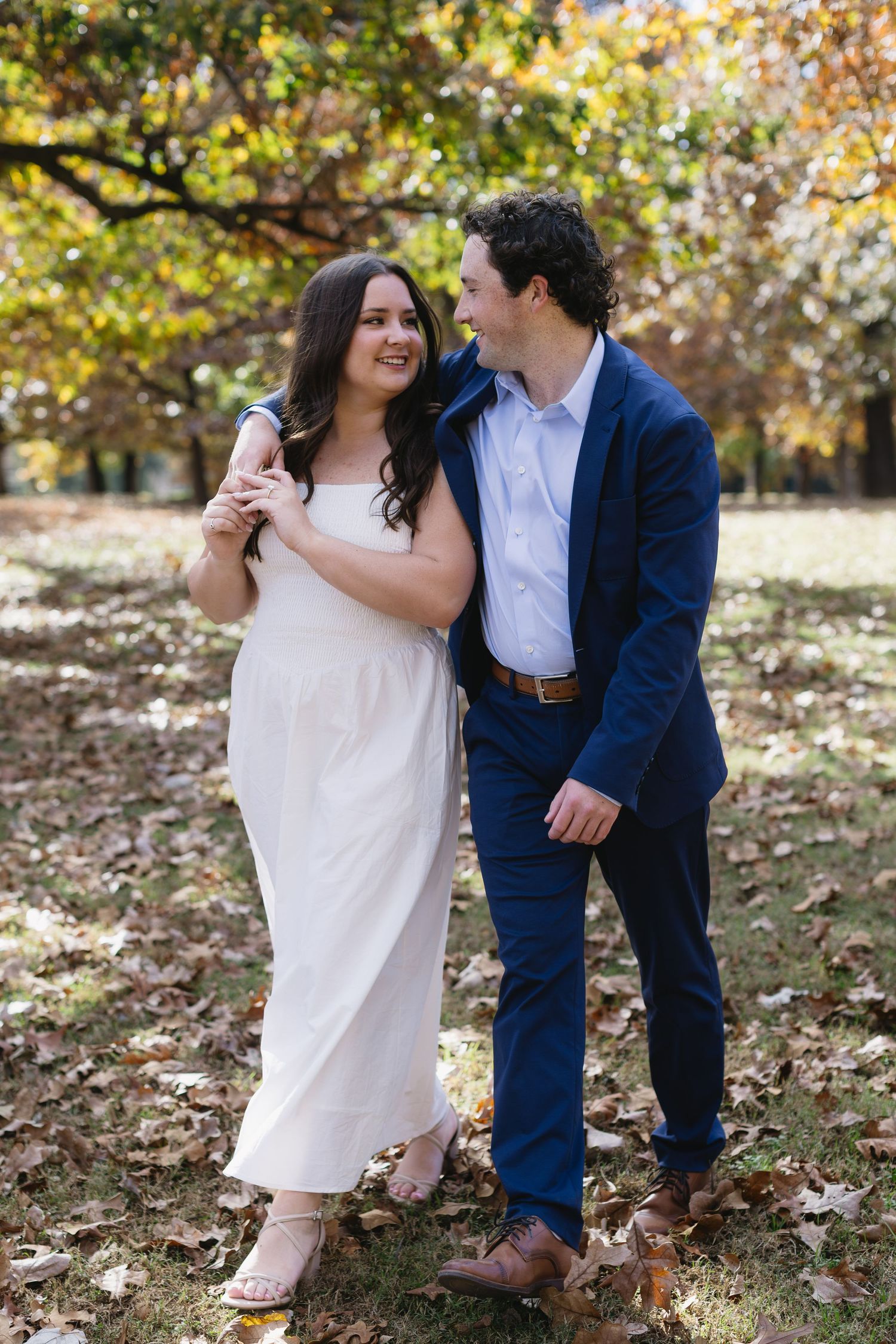 Couple in white dress and navy suit sharing playful moments among fall foliage.