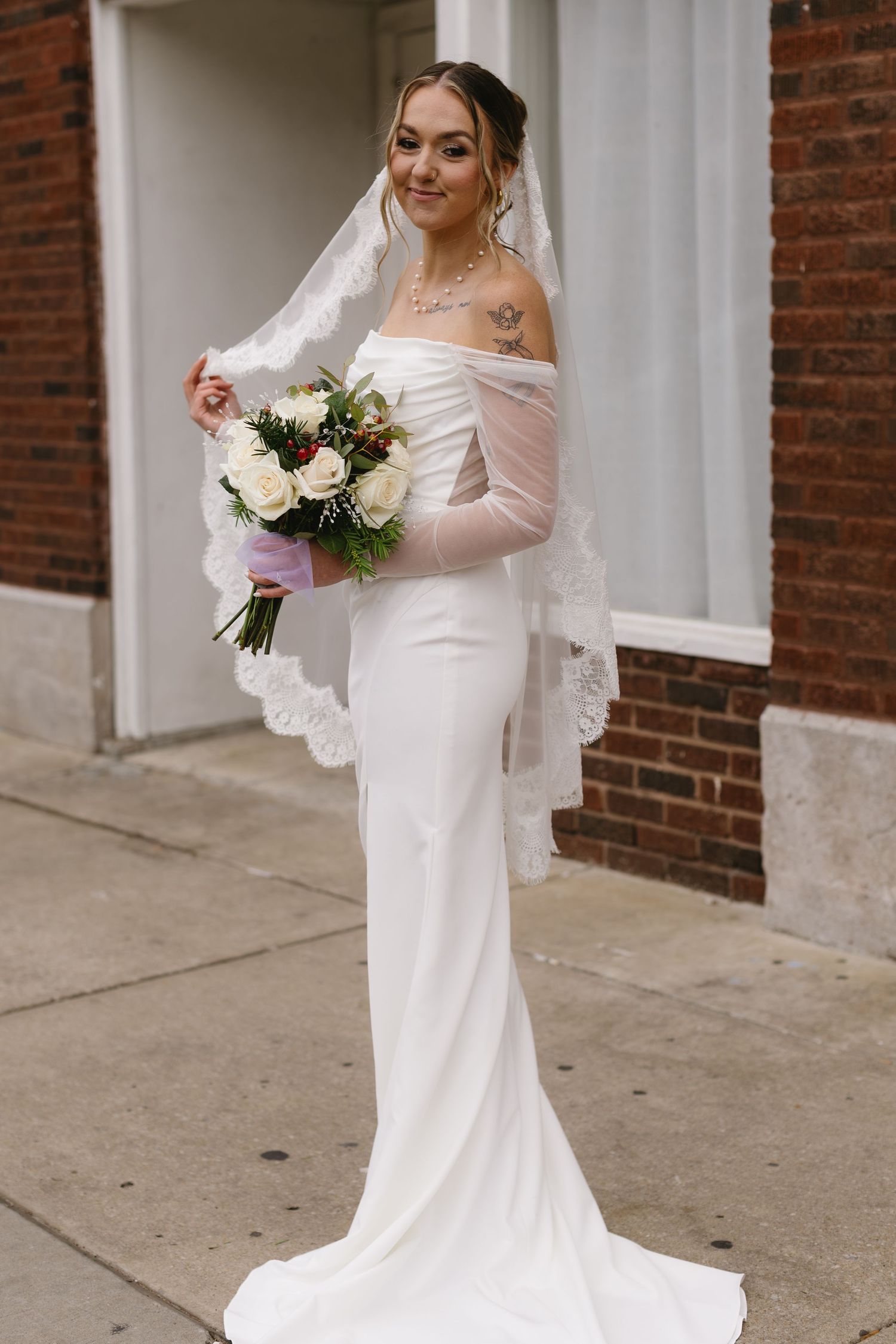 Elegant bride in long-sleeved white wedding gown and veil poses with bouquet against brick building.