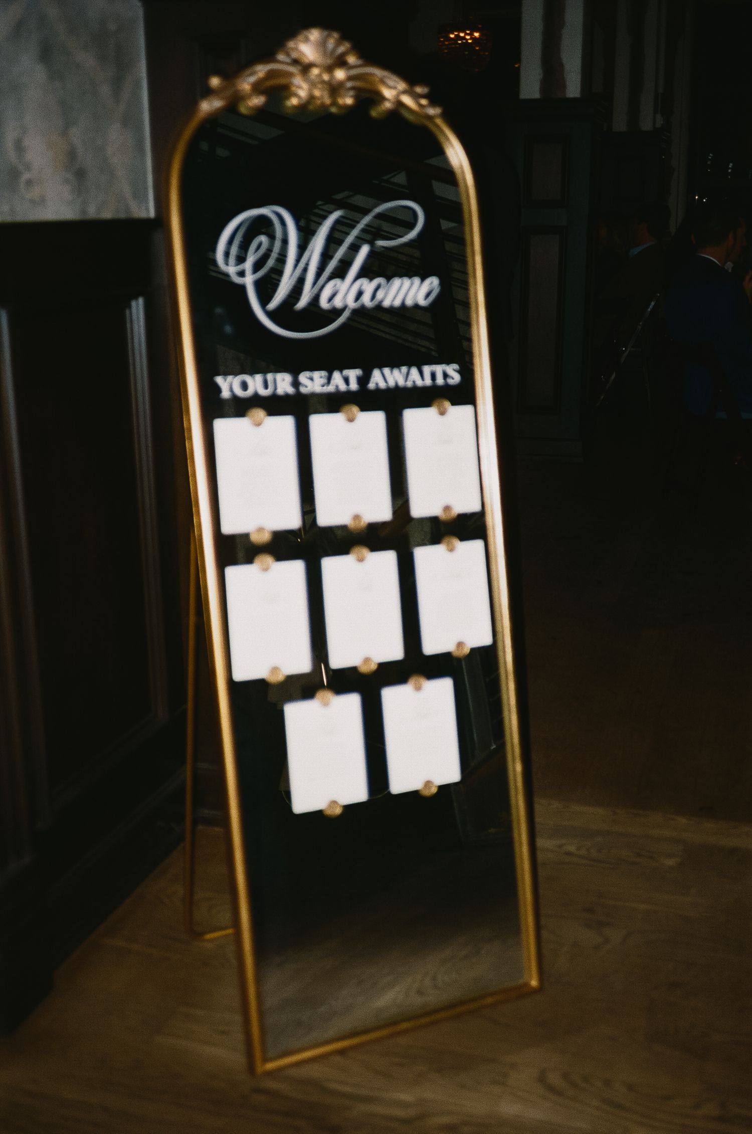 Elegant gold-framed seating chart display with 'Welcome' and 'Your Seat Awaits' text at a wedding reception.