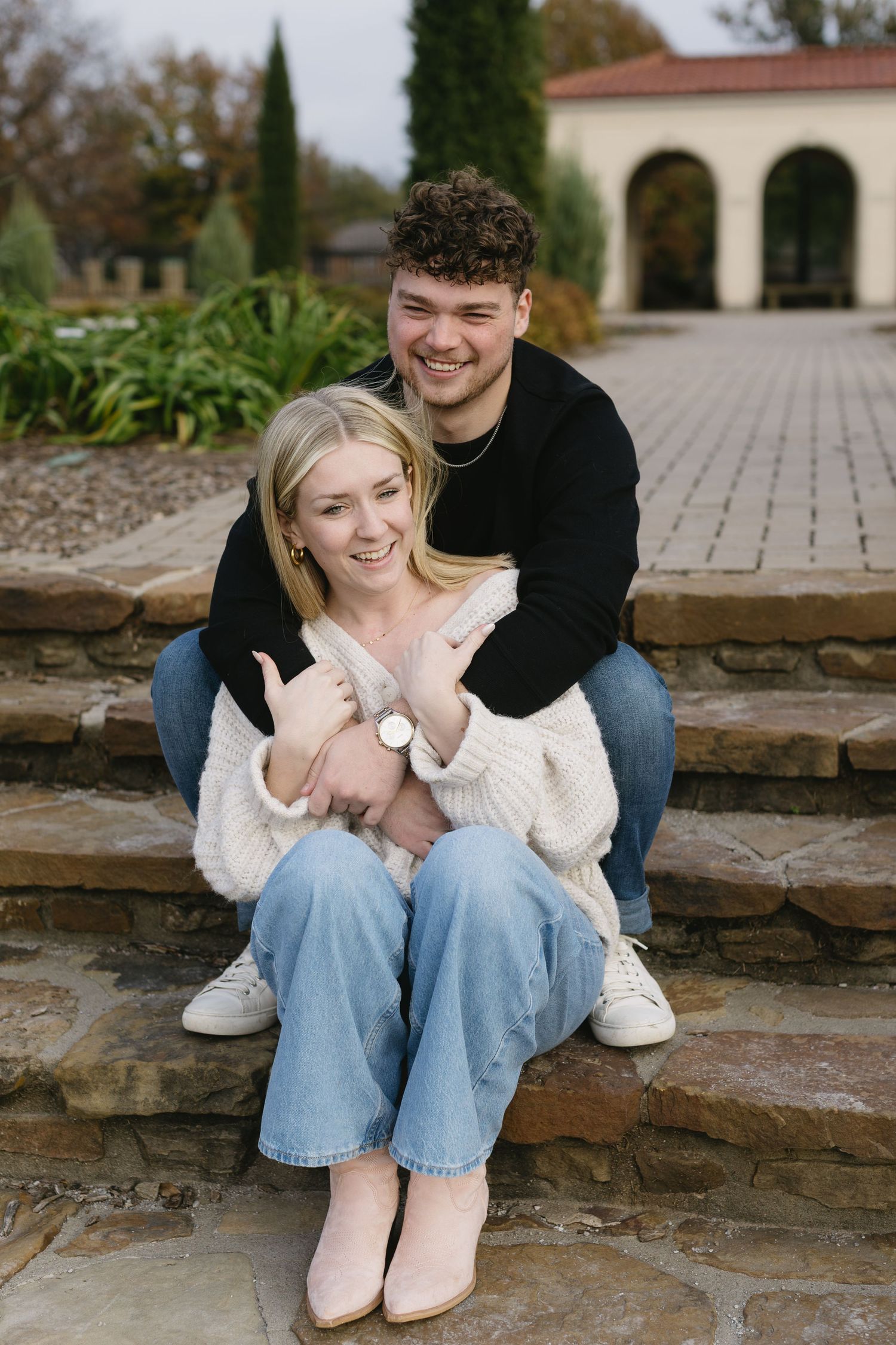 A couple sits together on stone steps, wearing casual sweaters and jeans on an autumn day.