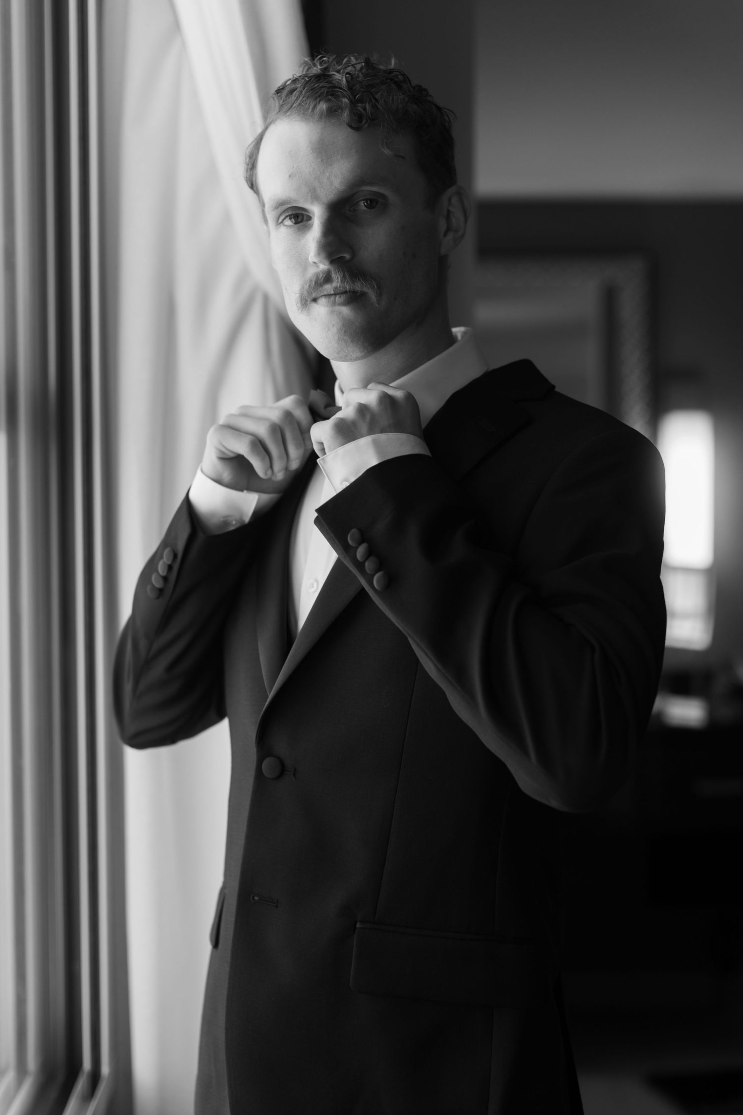 Silhouetted figure in formal attire stands pensively by a window in dramatic black and white lighting.