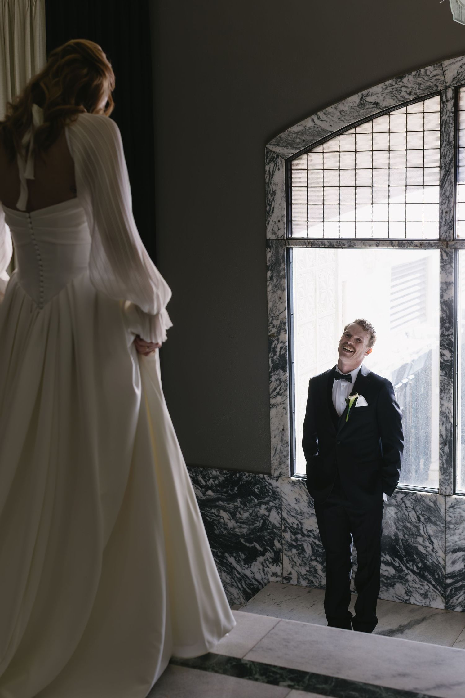 A romantic wedding first look moment captured in a stone-walled room with arched windows.