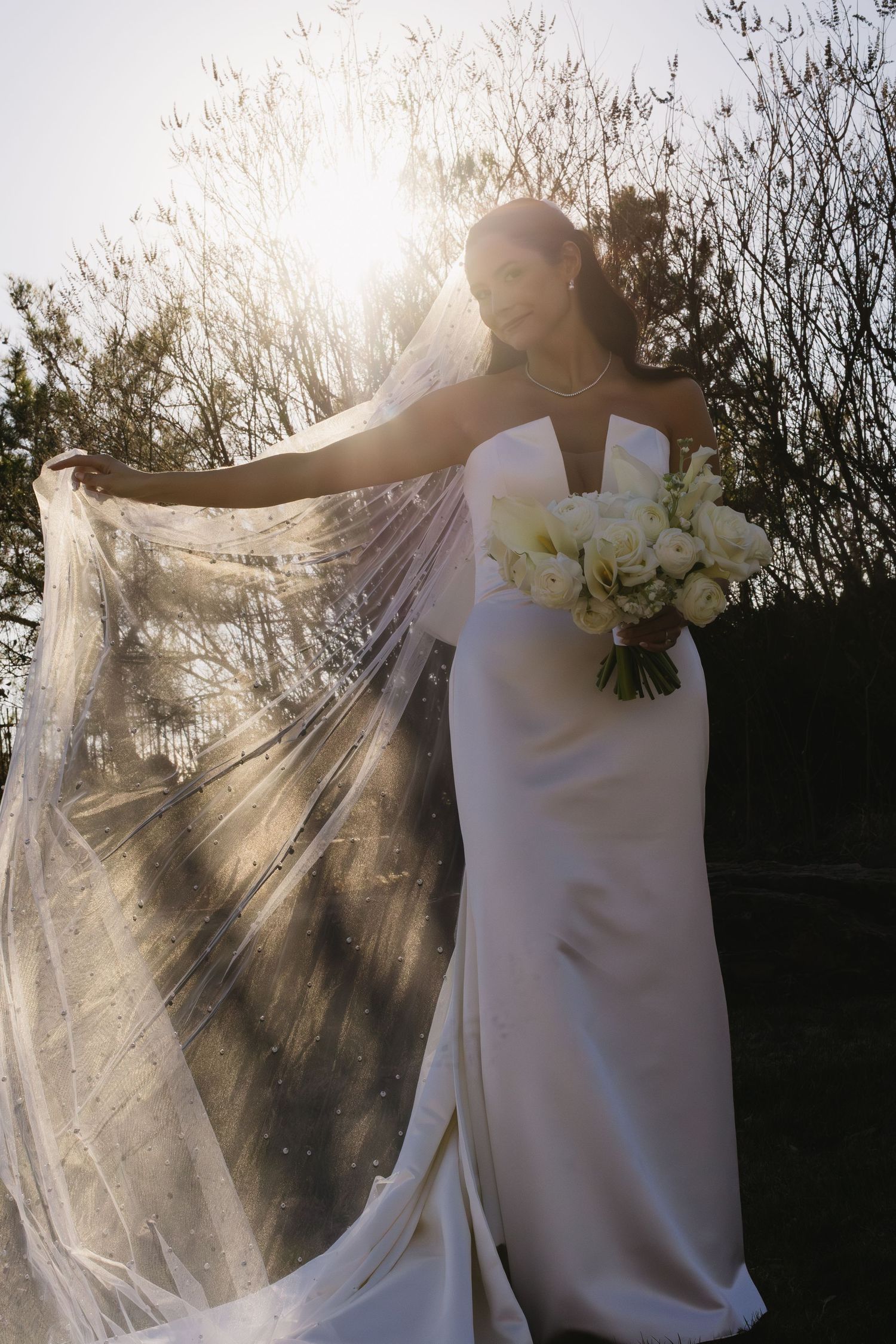 Sunlit bridal portraits showcasing a flowing veil and white bouquet against bare trees.
