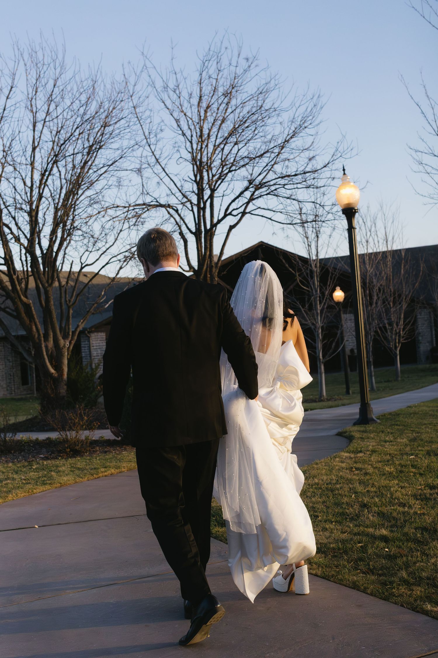 A newlywed couple walks along an illuminated path at dusk with vintage lampposts.