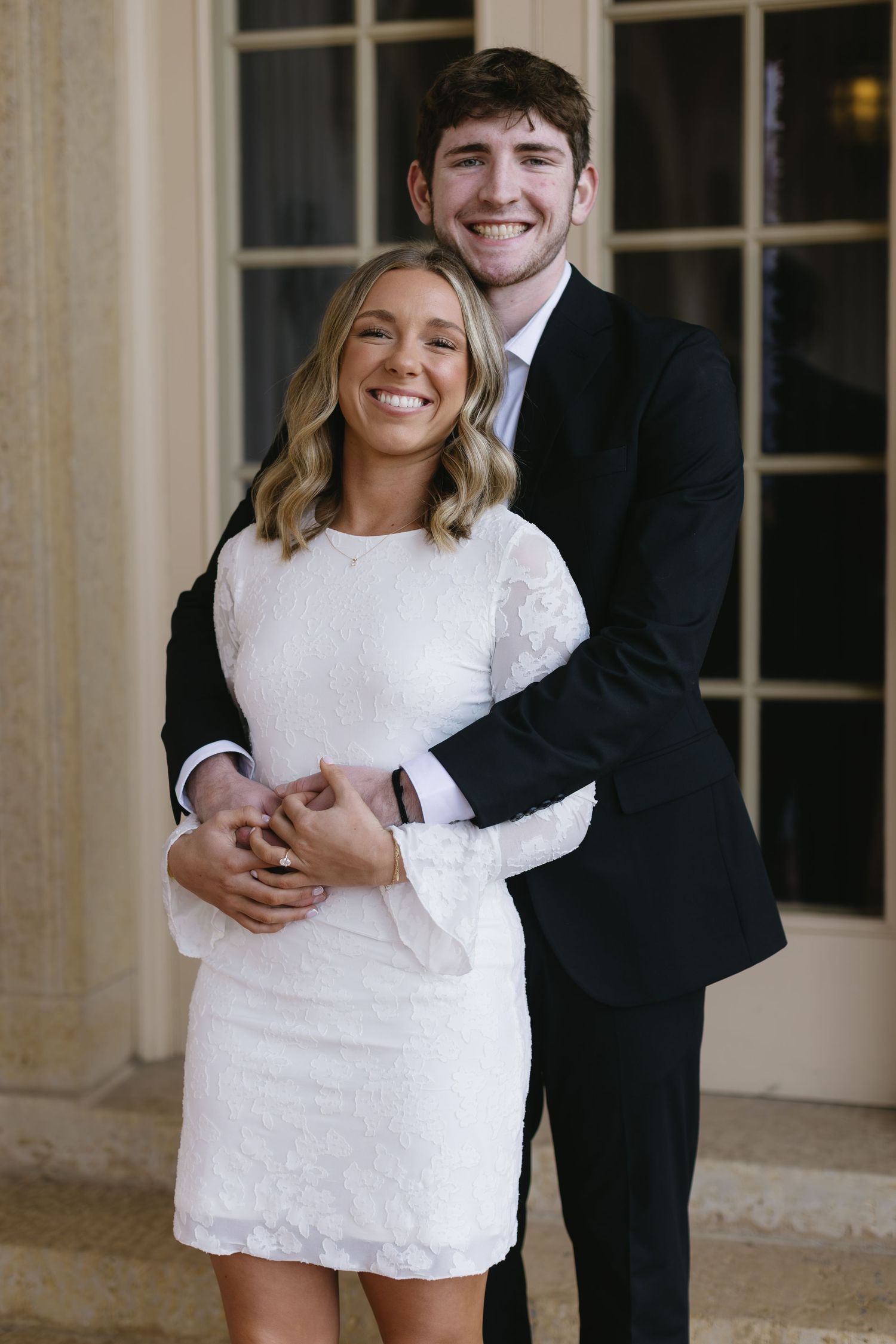A couple in formal attire embraces and smiles during a photo session with elegant windows in the background.