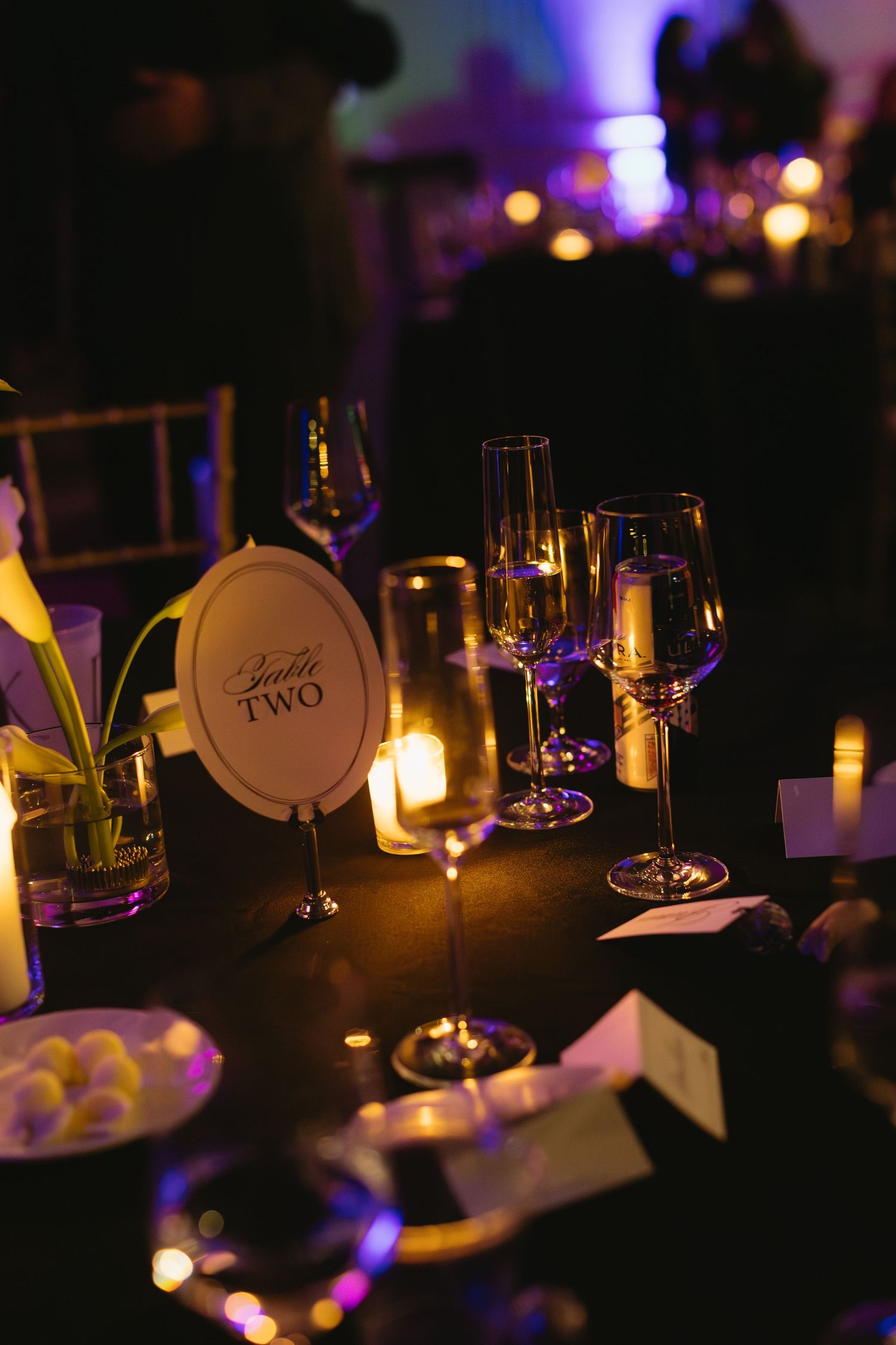 Candlelit wedding reception tables with wine glasses and table numbers in romantic lighting.