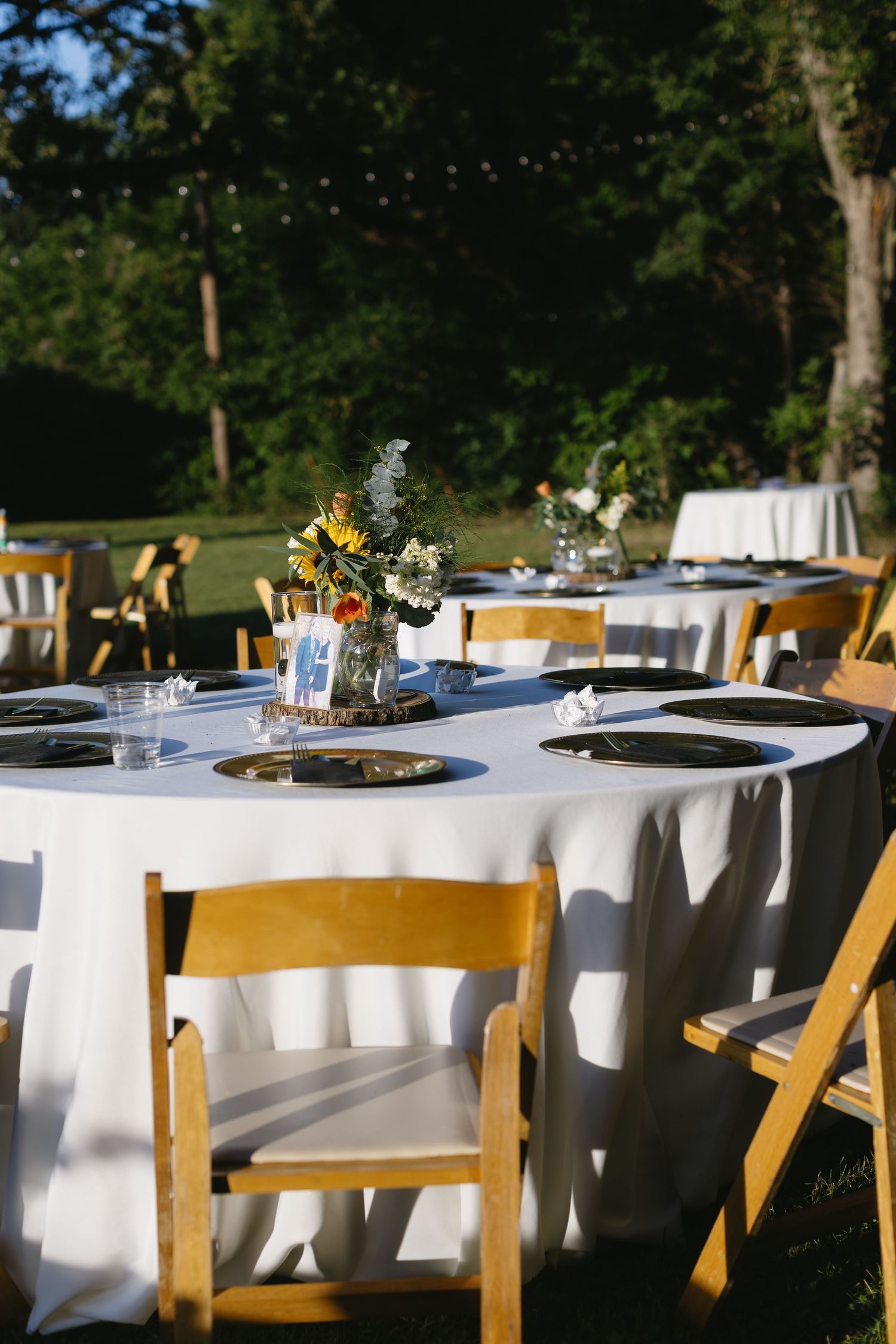 Elegant outdoor wedding reception tables set with white linens and wooden chairs under trees.