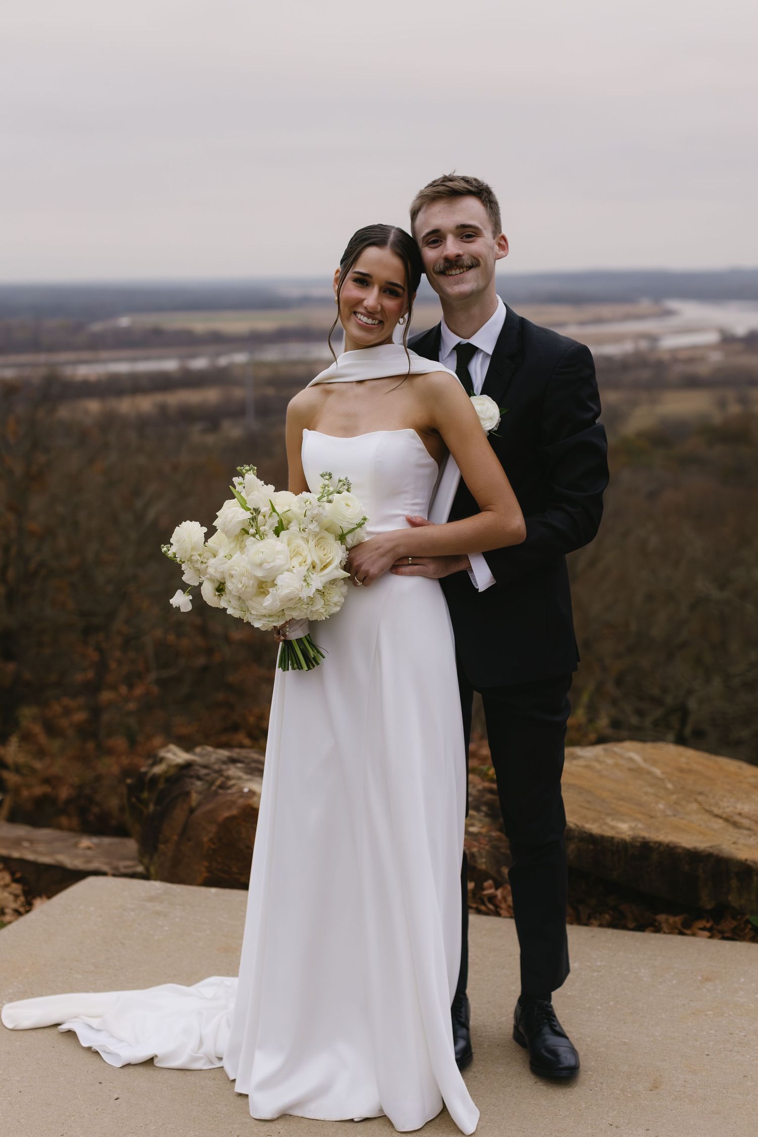 Couple in wedding attire embracing on scenic overlook with white bouquet.