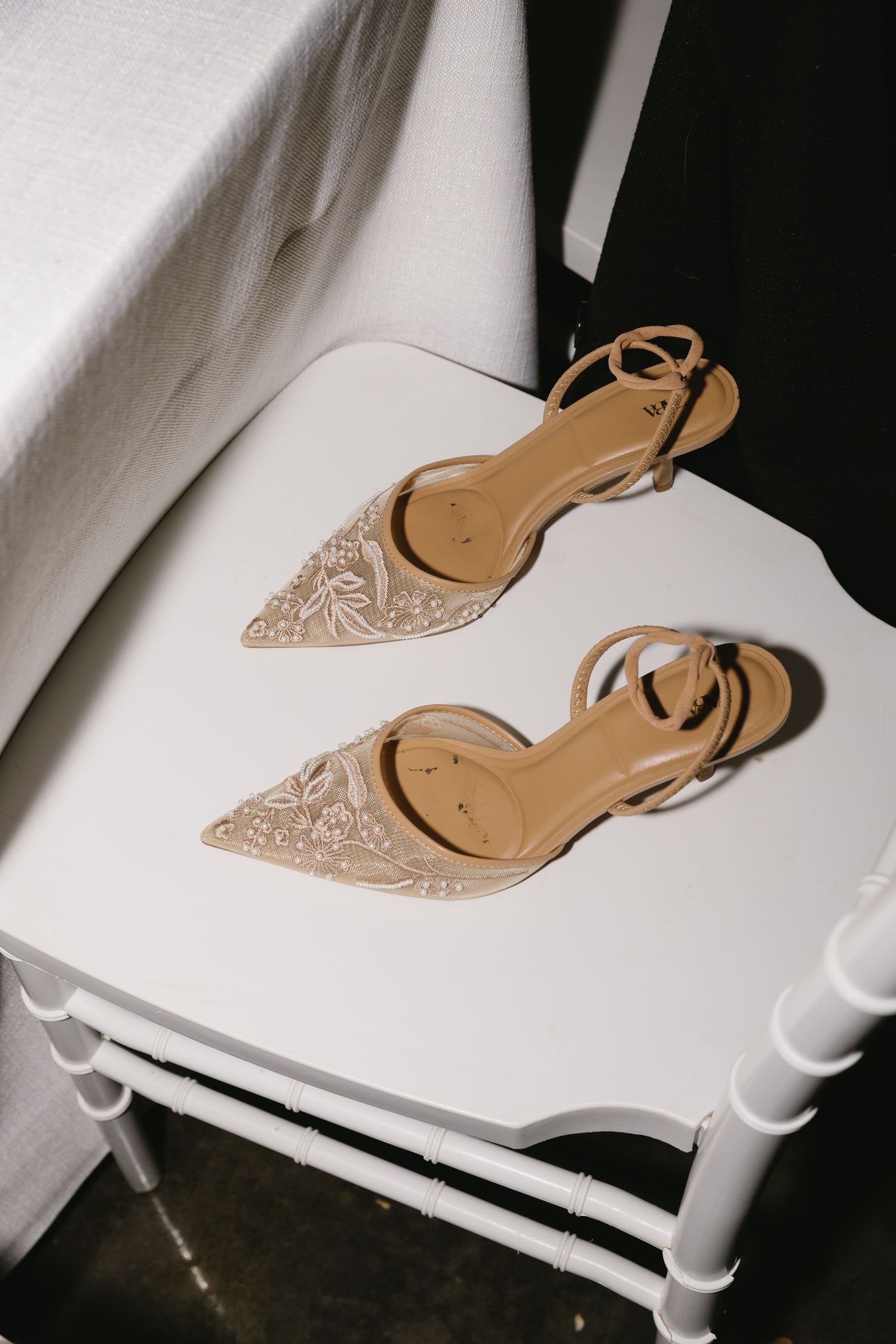 Delicate beige bridal shoes with intricate lace detail sitting on a white chair.