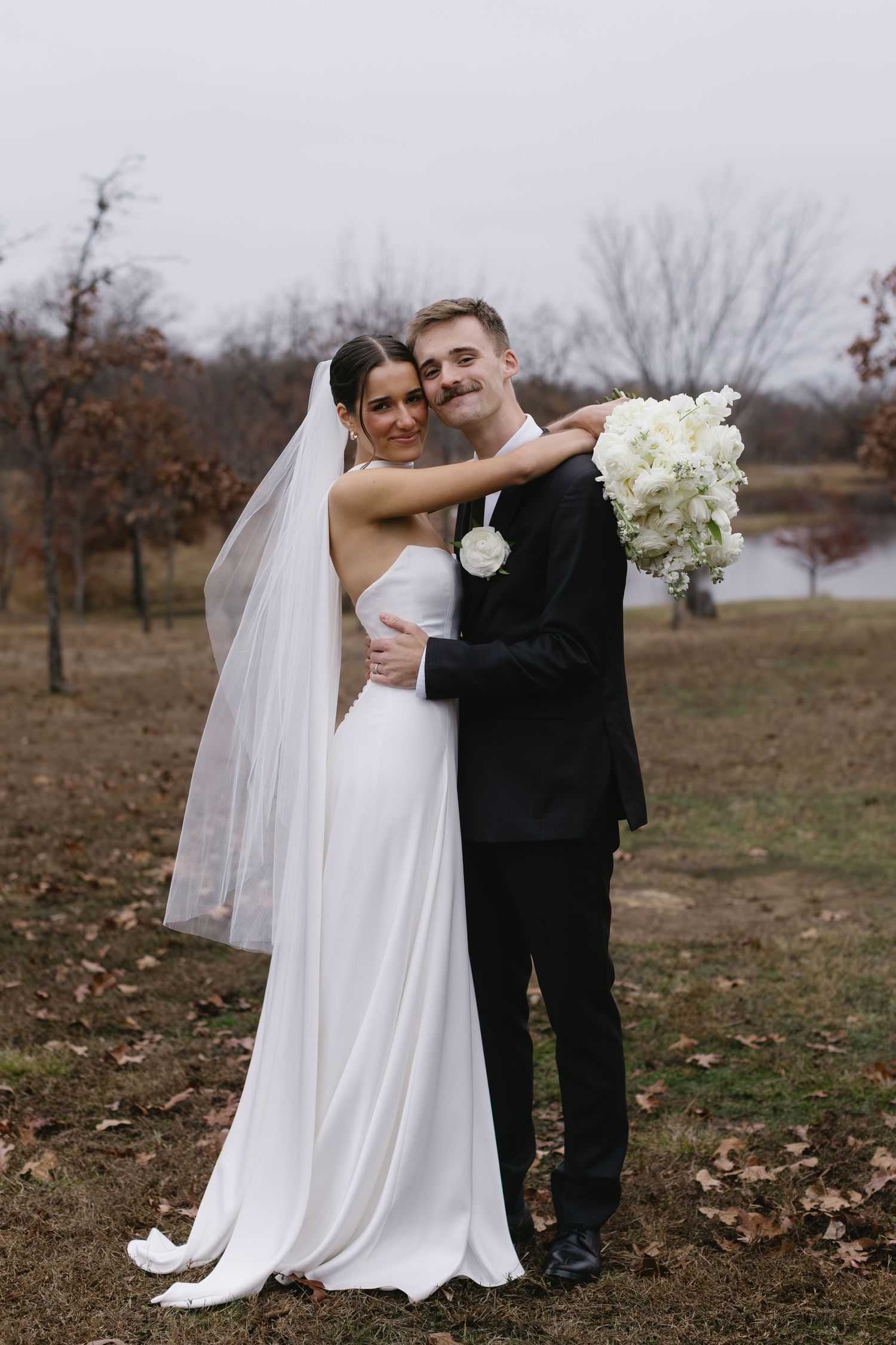 Wedding couple embracing in autumn field with white flowers and flowing veil.