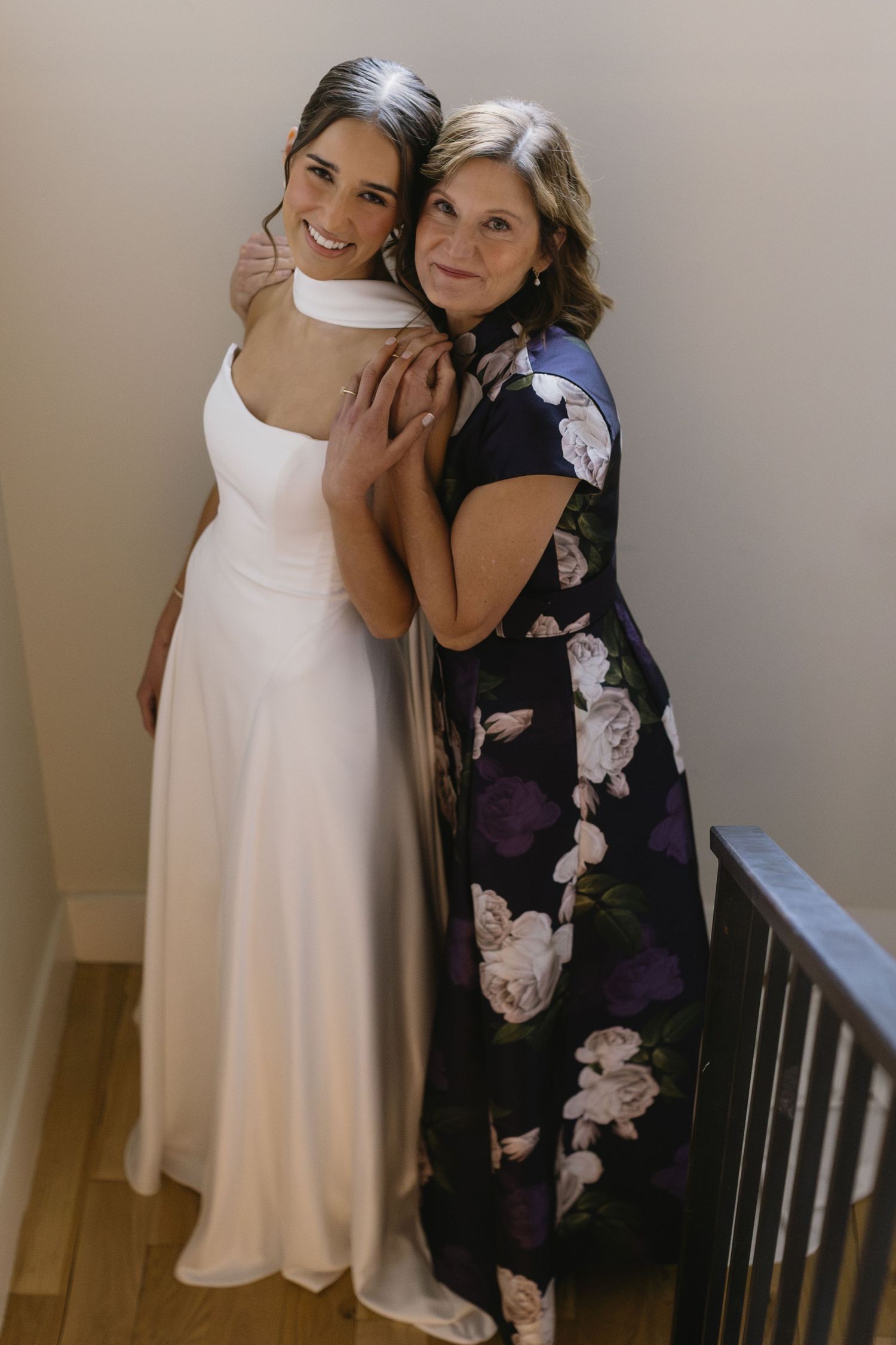 Two people embracing in formal attire, one wearing a white dress and the other in a floral navy dress.