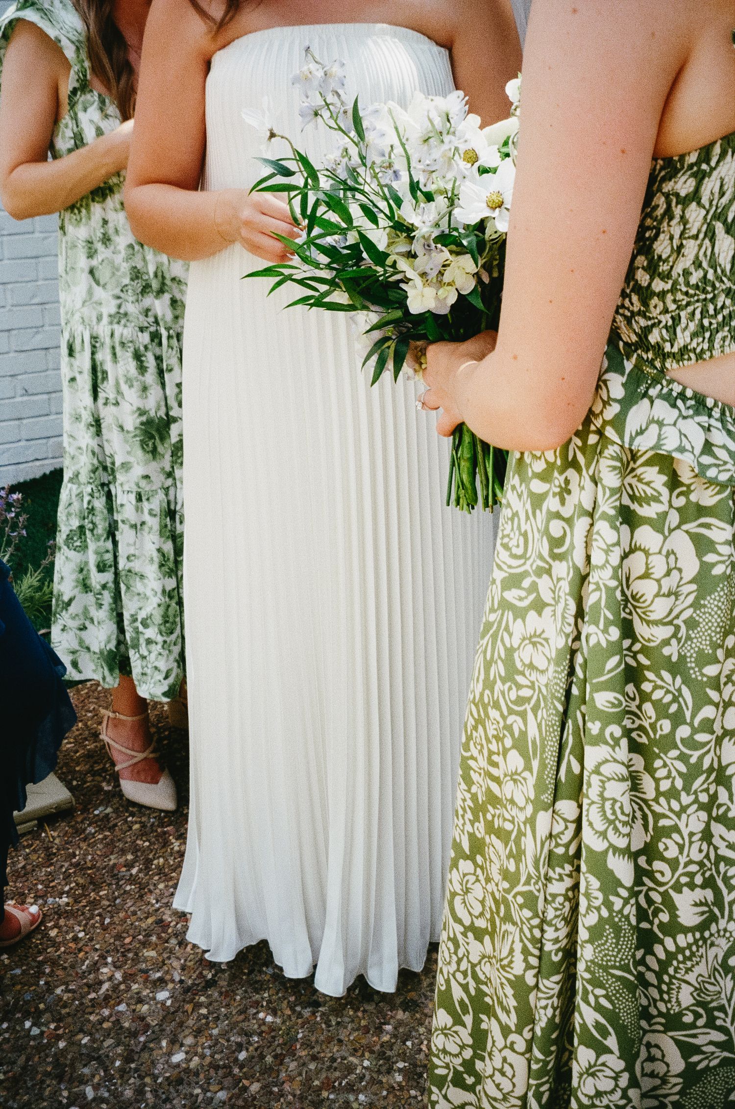 Wedding party wearing pleated white gown and green floral dresses holding white bouquet with greenery at outdoor ceremony.