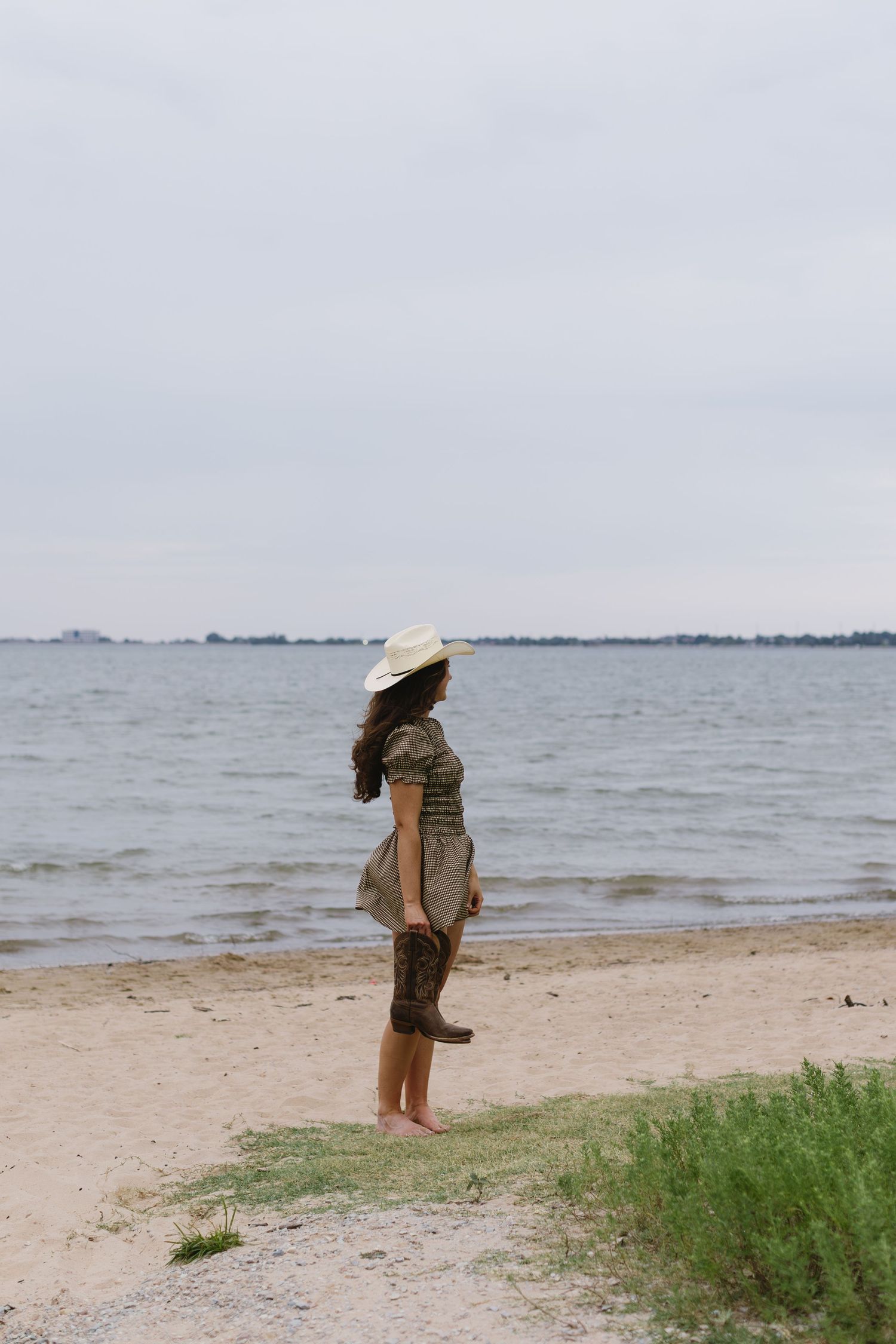 Person in casual clothing and hat standing alone on a peaceful beach shoreline.
